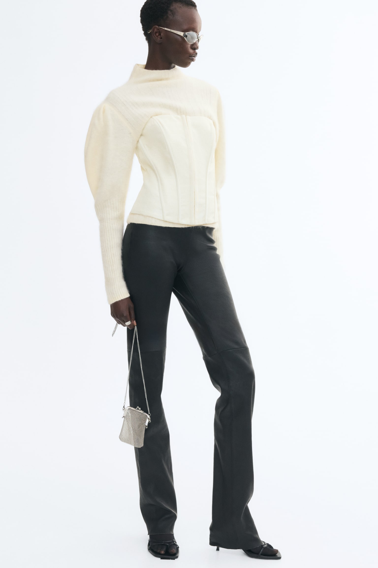 A fitted cream sweater in rib-knit wool, featuring a high neck and dramatic puff sleeves with long ribbed cuffs, is worn by the model. The sweater is layered under a matching structured cream corset and styled with black trousers. - 1