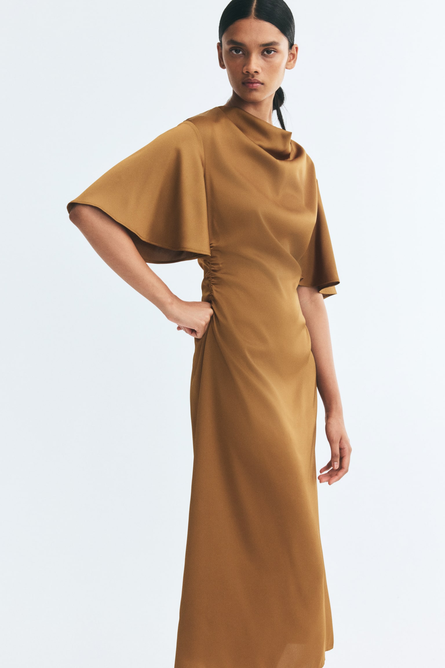 The model is wearing a calf-length, fitted satin dress in a golden-brown hue, standing with one hand resting on her waist. The dress features a draped cowl neckline, wide butterfly sleeves, and gathered detailing at the side seam to create a ruched effect. - 1
