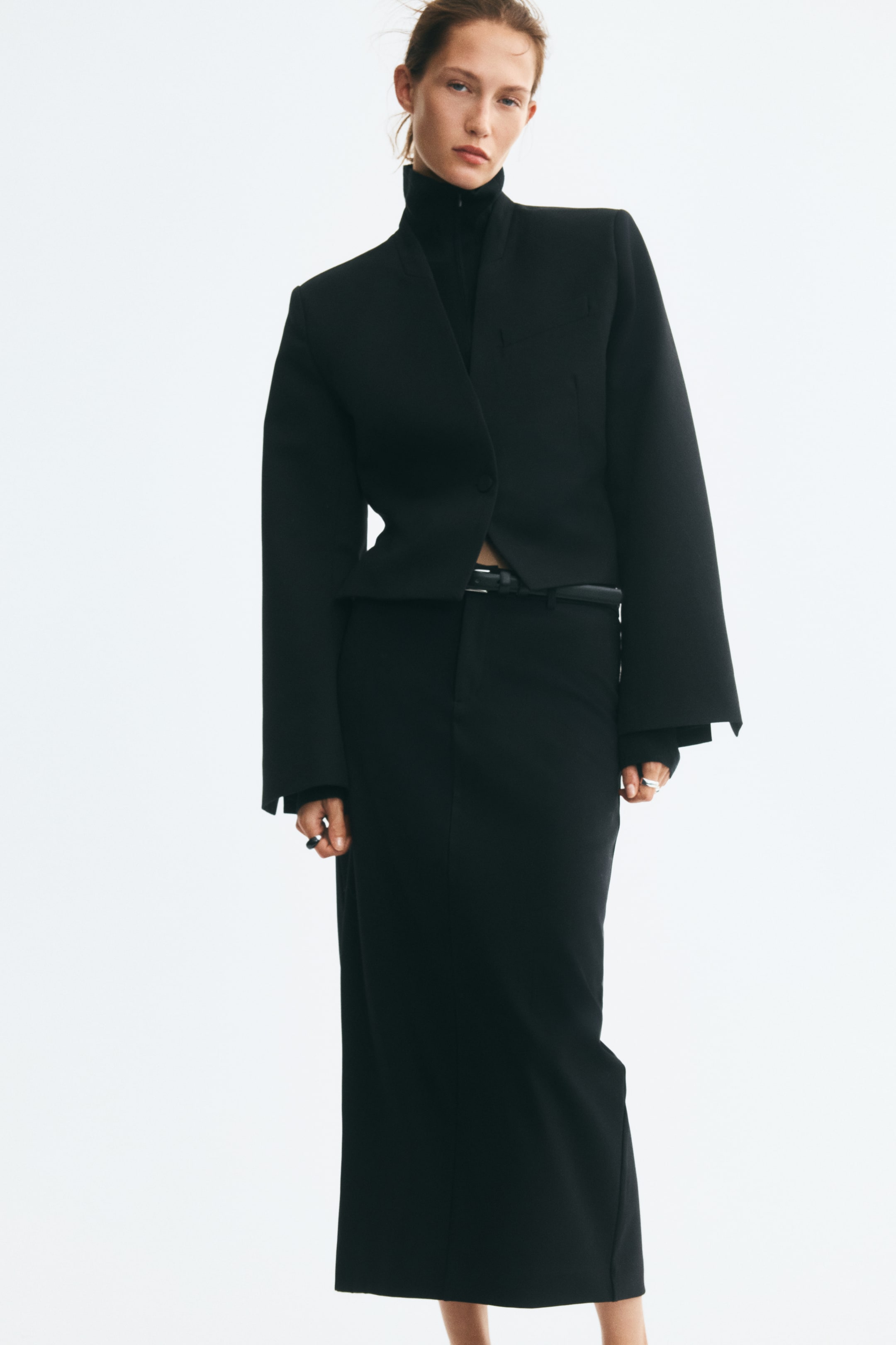 View larger image: The model is wearing a long black skirt in stretch twill, styled with a thin black belt at the waist. The high-waisted, straight-cut skirt is paired with a cropped black jacket and a black turtleneck.