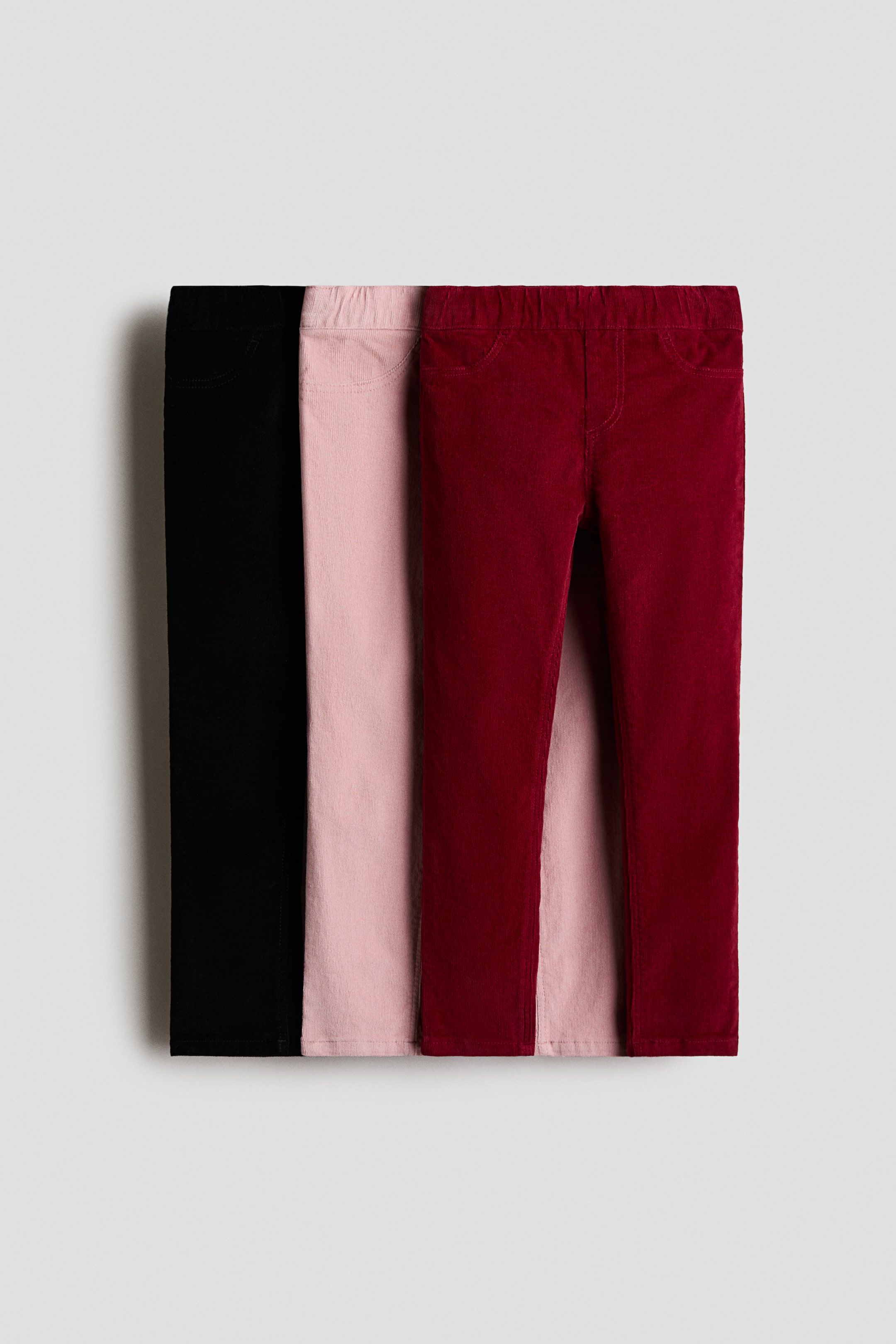 Ampliar la imagen: Tres pares de jeggings de pana en negro, rosa claro y rojo oscuro se superponen en una vista frontal, mostrando su textura de fina canalé. El par superior color borgoña presenta una cintura elástica, costuras simuladas de bragueta y bolsillos frontales simulados.
