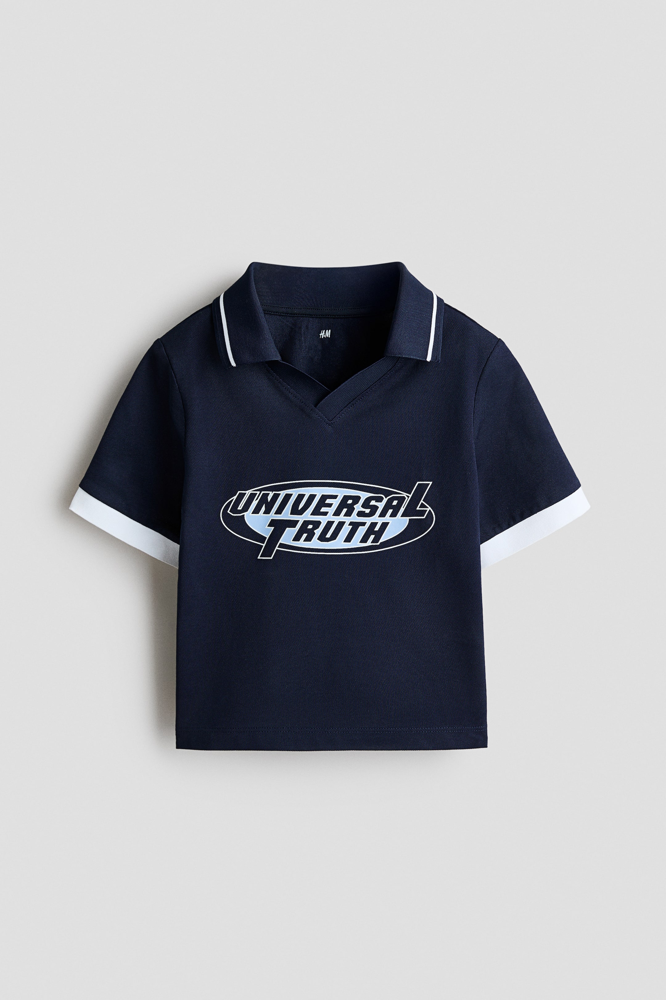 Xem ảnh lớn hơn: Áo polo xanh navy dáng ngắn đang hướng mặt trước, nổi bật với cổ áo nhọn viền trắng và phần mở cổ chữ V. Áo có tay ngắn với viền trắng và họa tiết in đồ họa trung tâm phía trước, hiển thị dòng chữ 'UNIVERSAL TRUTH' trong thiết kế hình oval.