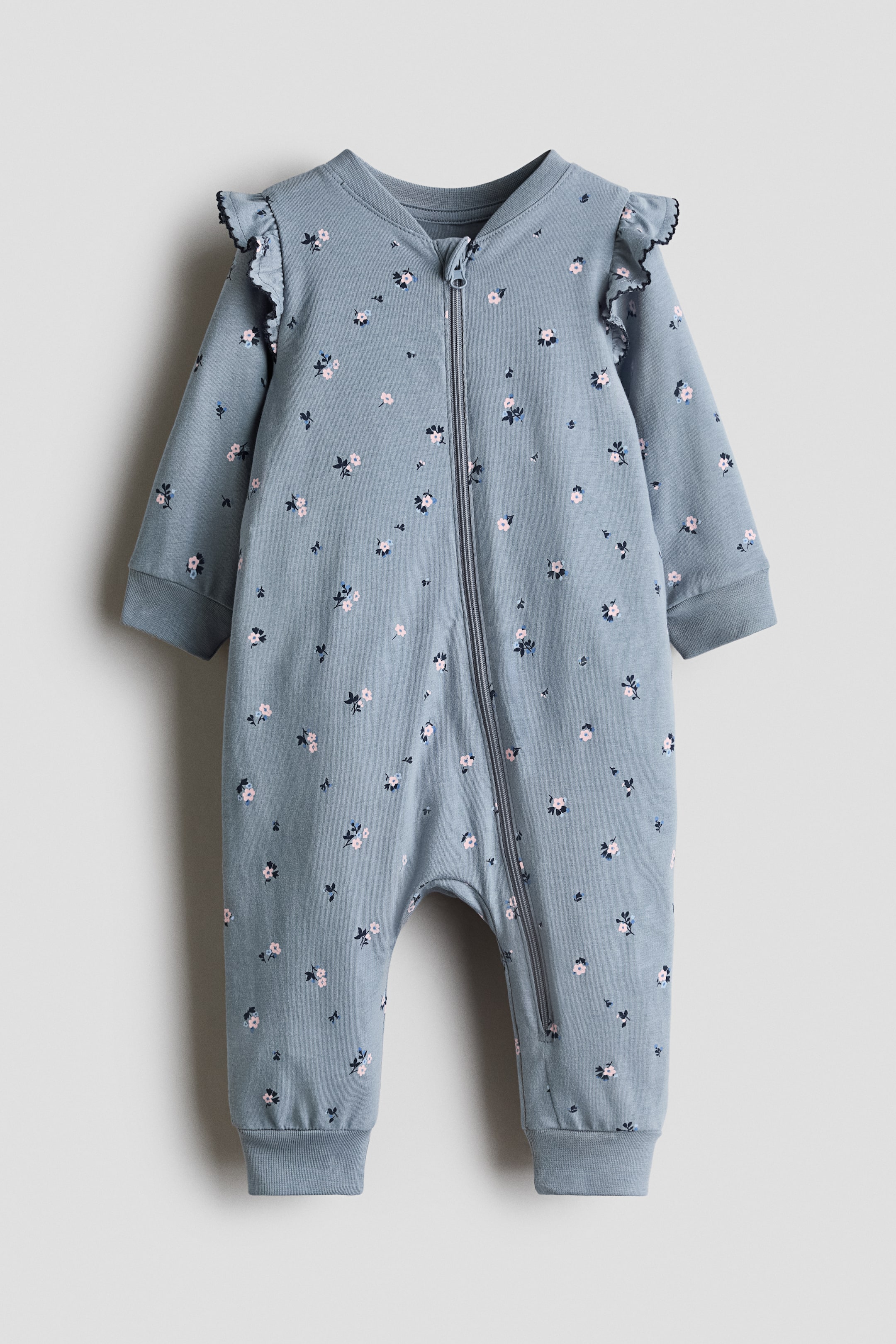 Visa större bild: En sleepsuit visas framifrån, tillverkad av mjuk, blågrå tryckt bomullsjersey med små blommönster i rosa och blått. Den är designad med dekorativ volangkant över axlarna, V-ringning, dragkedja som sträcker sig längs framsidan och ner längs ett ben samt muddliknande avslut vid handleder och fotleder.