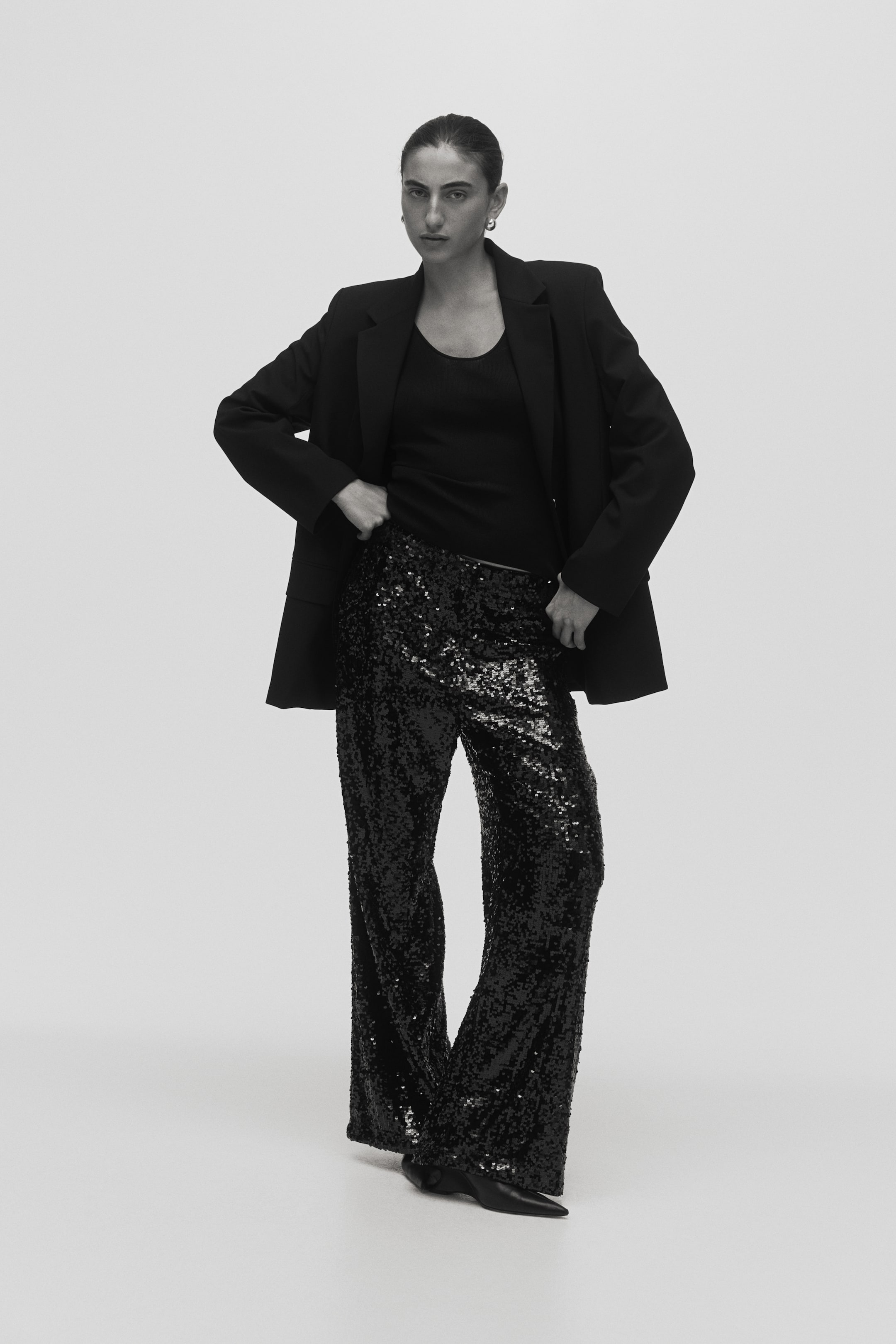 Visa större bild: Svarta paljettbyxor visar upp en skimrande textur och en bred benklippning. De presenteras från en frontvänd vinkel, sträcker sig till golvet och bildar en liten pöl vid anklarna och är stylade med en svart topp och en överdimensionerad svart blazer.