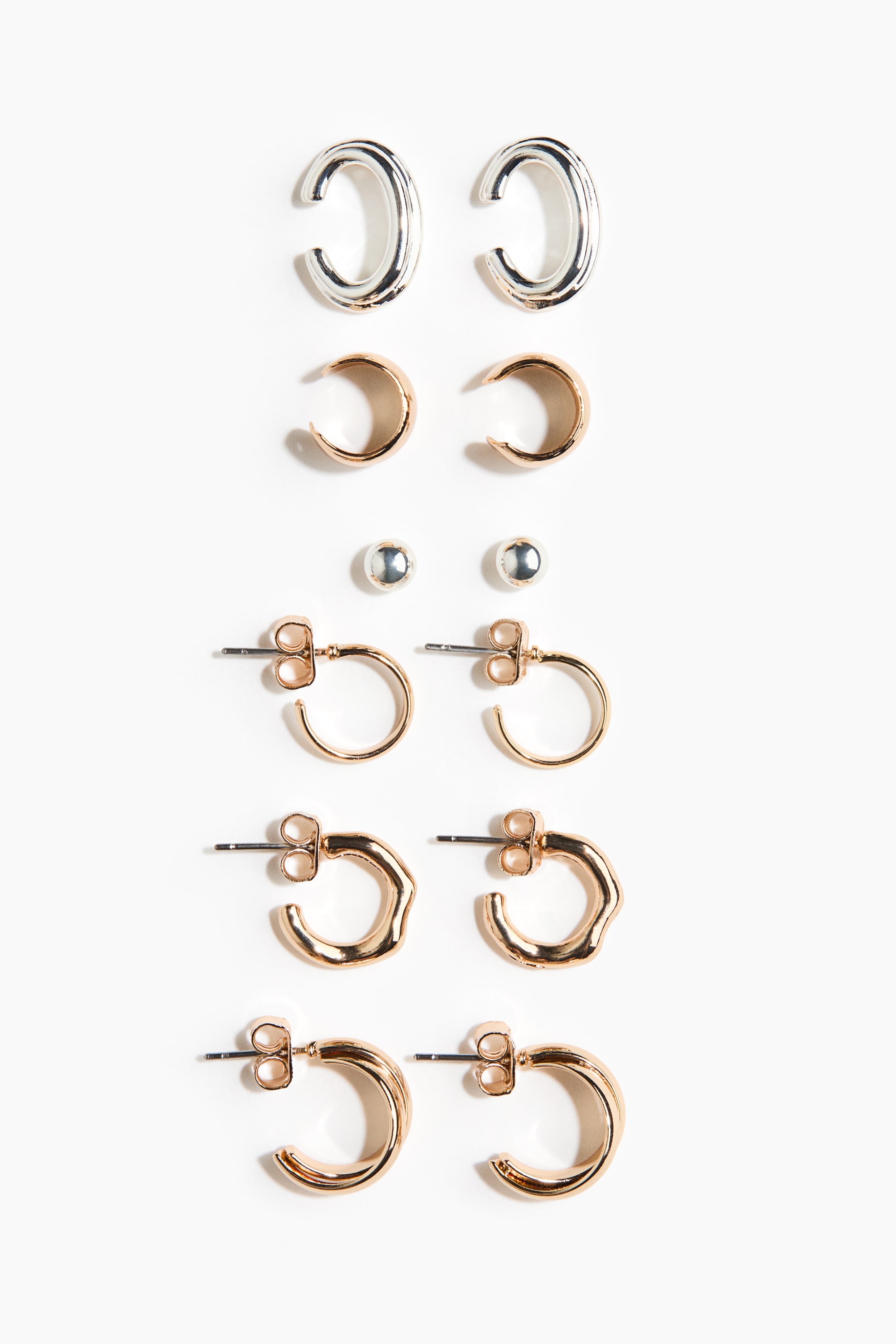 View larger image: An earring set featuring six pairs of earrings presents a mix of silver-tone and rose gold-tone designs. The collection showcases a range of styles arranged vertically, including textured C-shaped hoops, wide smooth hoops, small ball studs, thin classic hoops, wavy-edged hoops, and double-layered hoops.