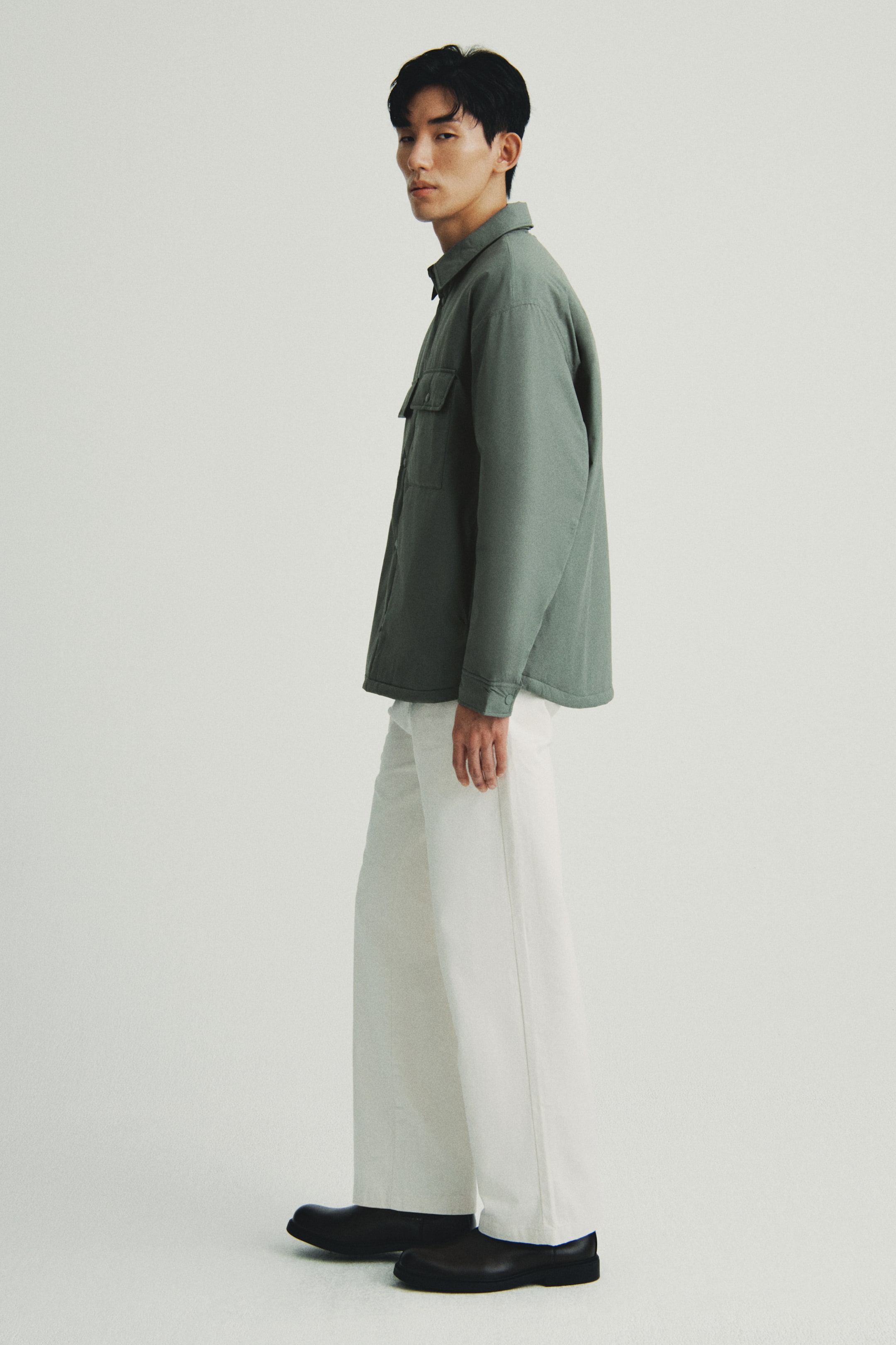 이미지 확대 보기: 한 사람은 좌측을 향해 있는 상태에서 카라, 두 개의 가슴 플랩 포켓, 그리고 단추로 잠글 수 있는 소맷단이 있는 차분한 녹색 셔켓을 입고 있습니다. 이 셔켓은 아이보리색 와이드 레그 트라우저 위에 레이어드되어 있으며, 두꺼운 밑창을 가진 검정 슬립온 신발로 전체적인 옷차림을 완성합니다.