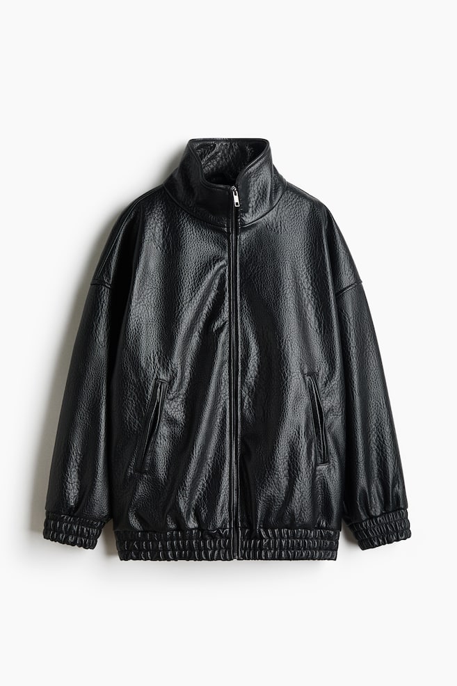 A black, textured-leather jacket is facing the front, featuring a high stand-up collar and a full-length zipper closure. The jacket is showcasing dropped shoulders, side welt pockets, and elasticated, ruched detailing at the cuffs and hem. - 1