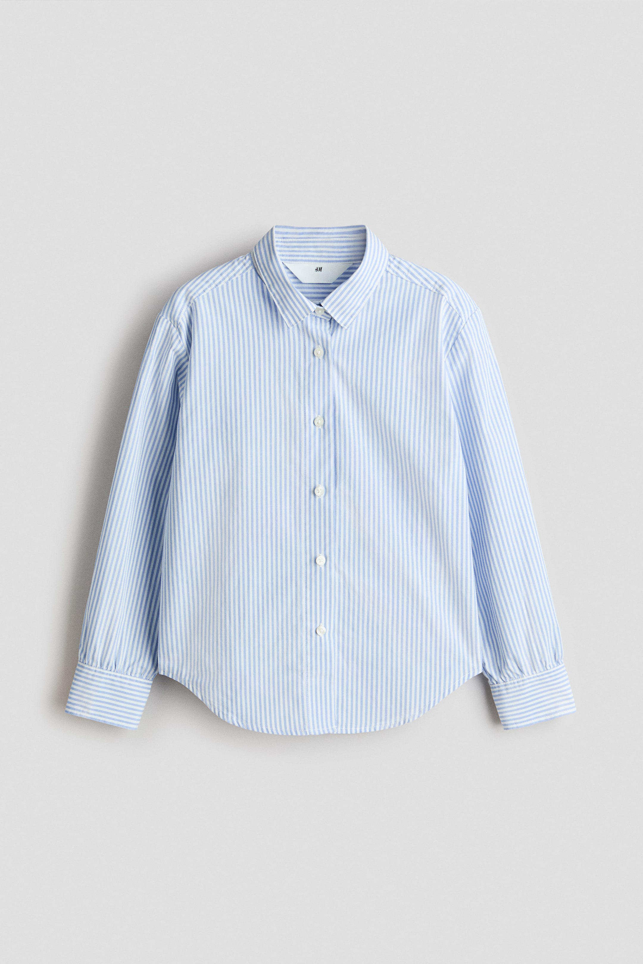 Agrandir l'image: Une chemise à rayures bleu clair et blanche est posée à plat, montrant le devant. Elle présente un col pointu, une patte de boutonnage sur le devant avec des boutons blancs, des manches longues munies de poignets rayés et boutonnés, et un ourlet arrondi.