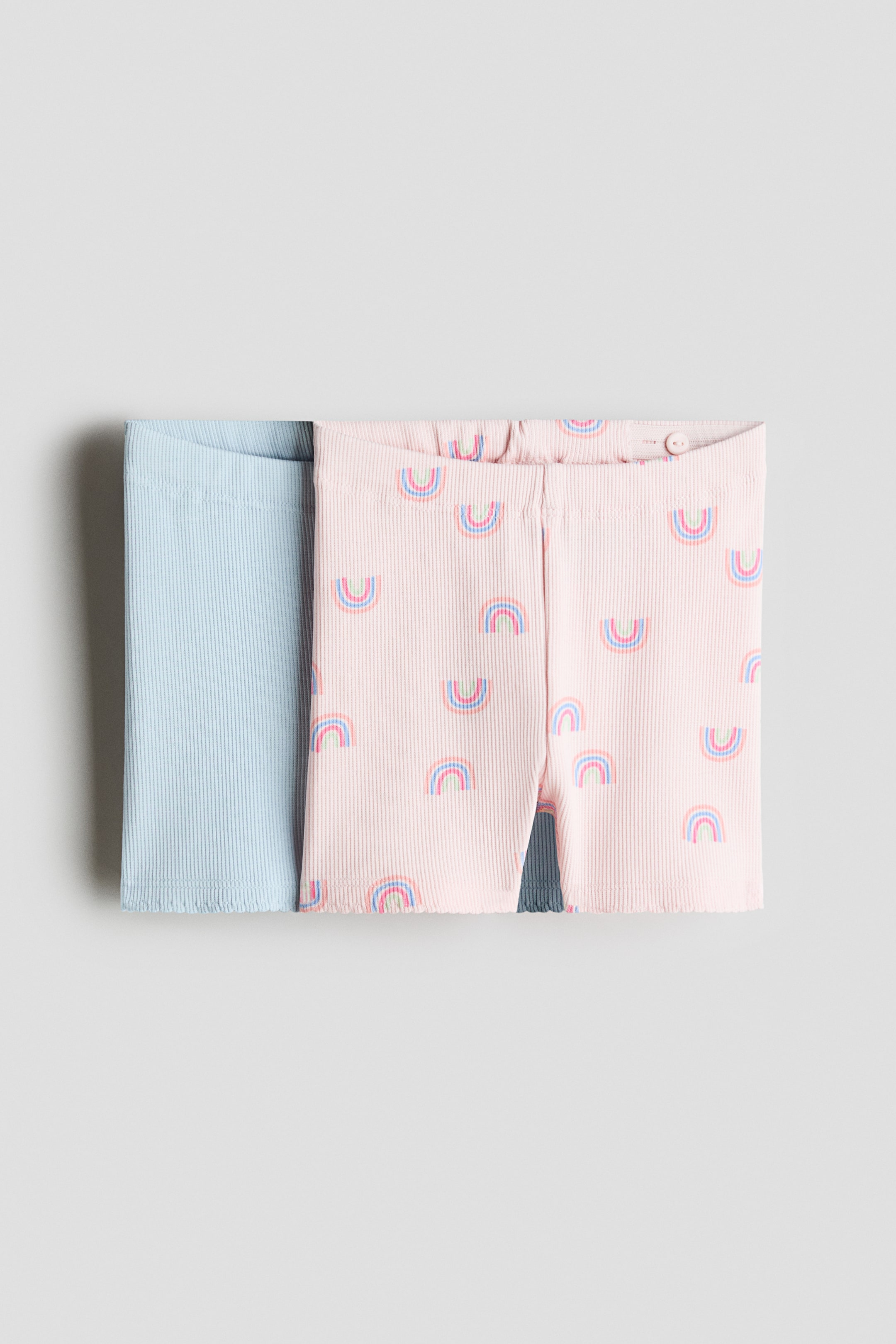 Ingrandisci l'immagine: Due paia di pantaloncini ciclistici a coste sono presentati nella vista frontale. Il paio rosa chiaro presenta piccoli motivi arcobaleno, una vita elasticizzata regolabile e orli smerlati. Il paio azzurro solido mostra orli smerlati.