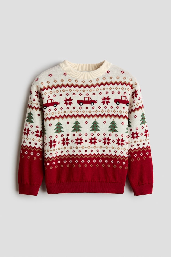 A jacquard-knit jumper is shown from the front, featuring a cream and red Fair Isle-style pattern with horizontal bands of red pick-up trucks, green pine trees, and geometric snowflake motifs. The jumper is designed with a cream-coloured ribbed round neckline, dropped shoulders, and long sleeves ending in red ribbed cuffs that match the ribbed hem. - 1