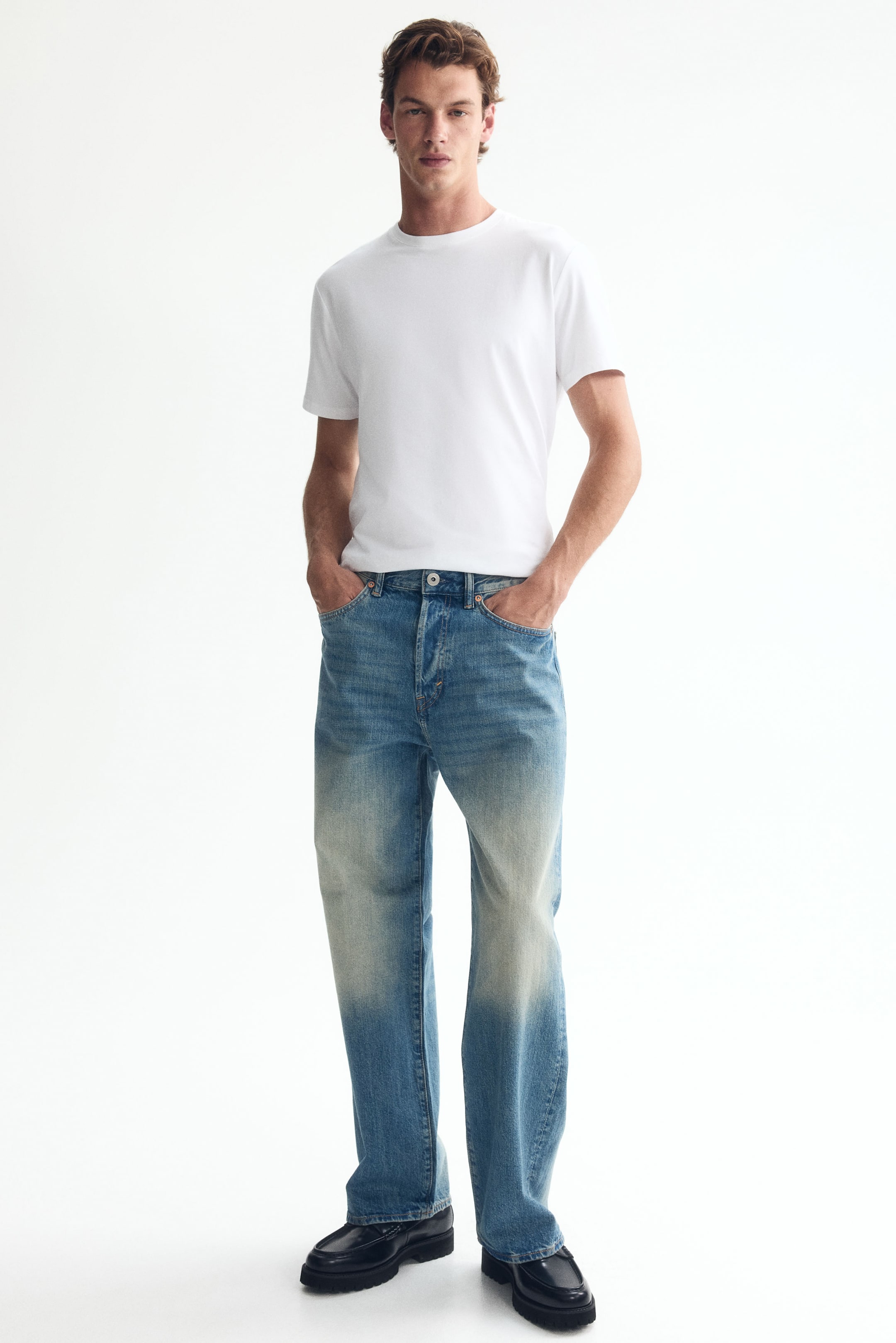 Voir une image plus grande: Le mannequin porte un jean à 5 poches en denim de coton rigide bleu clair avec un délavage usé, mettant en valeur une coupe ample à jambes larges avec un entrejambe bas et une taille régulière. Le jean est doté d'une fermeture à glissière, et le mannequin place ses mains dans les poches avant.