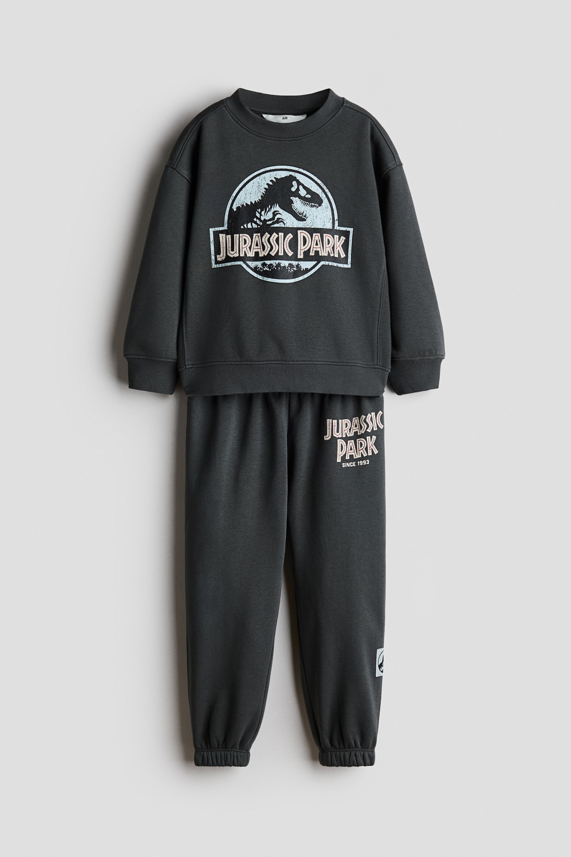 View larger image: A dark grey two-piece sweat set is shown from the front, featuring a long-sleeved sweatshirt with a round neckline, ribbed trim, and a large circular "Jurassic Park" graphic on the chest. The matching track pants have elasticated cuffs and a smaller "Jurassic Park" text graphic printed on the upper left leg.