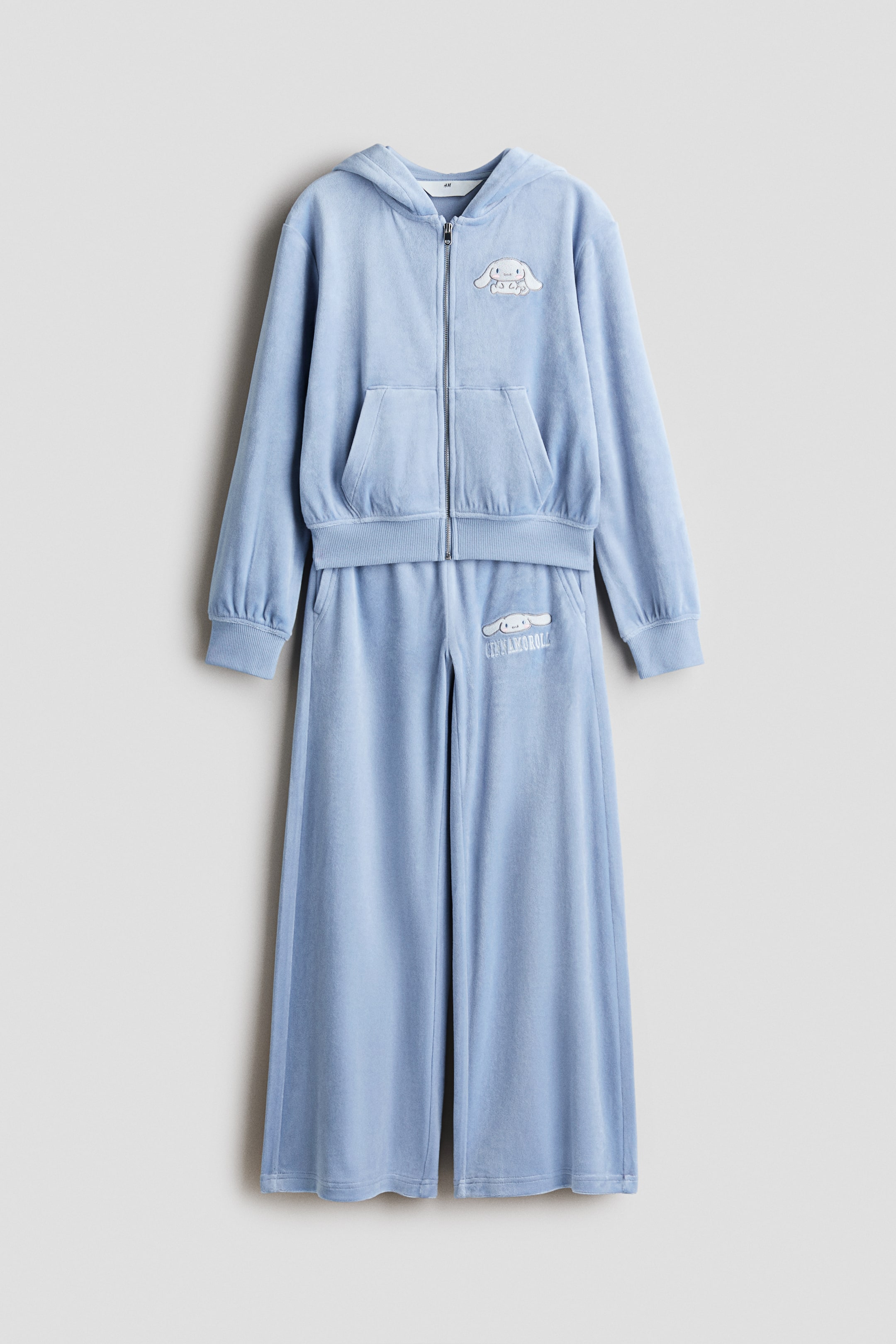 View larger image: A light blue velour two-piece set is laid flat and front-facing, featuring a full-zip hooded top with a kangaroo pocket, ribbed cuffs, and a ribbed hem. The top features a white embroidered Cinnamoroll character on the left chest, while the matching wide-leg pants feature a Cinnamoroll character and the text ‘CINNAMOROLL’ on the left thigh.