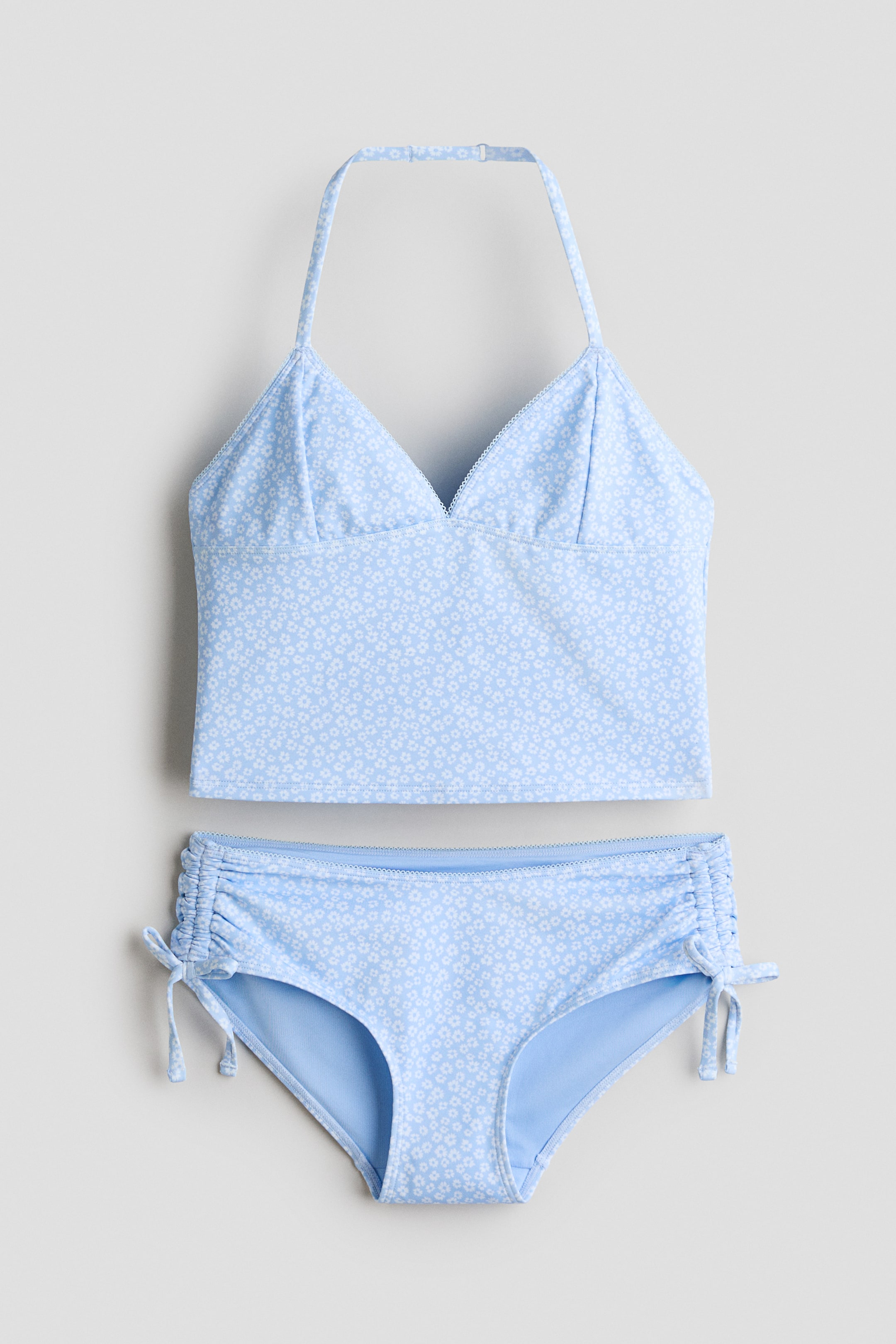 View larger image: A fully lined tankini set is showcasing its front, featuring a light blue top and matching bottoms adorned with a white floral print and picot trims. The tankini top boasts a V-neck and a slender, adjustable halterneck strap, while the bottoms are embellished with decorative drawstrings and sewn-in bows at the sides.