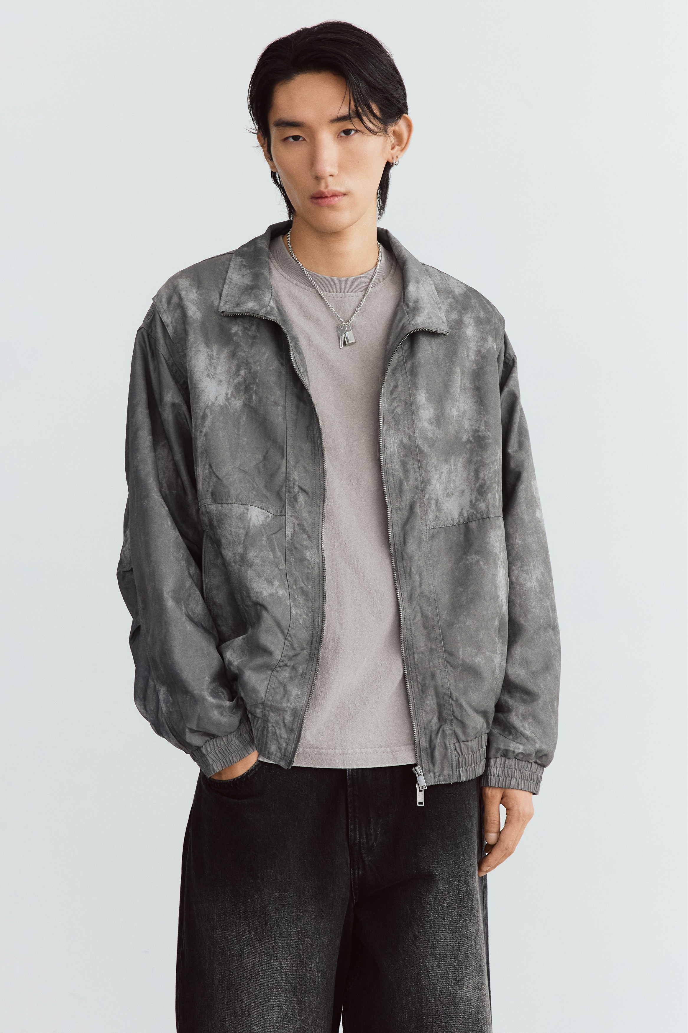 A grey, washed-effect zip-up jacket is displayed flat and facing forward, featuring a high stand-up collar, a central zip closure, and elasticated cuffs and hem. The jacket features vertical zippered side pockets and a textured, distressed finish throughout.
