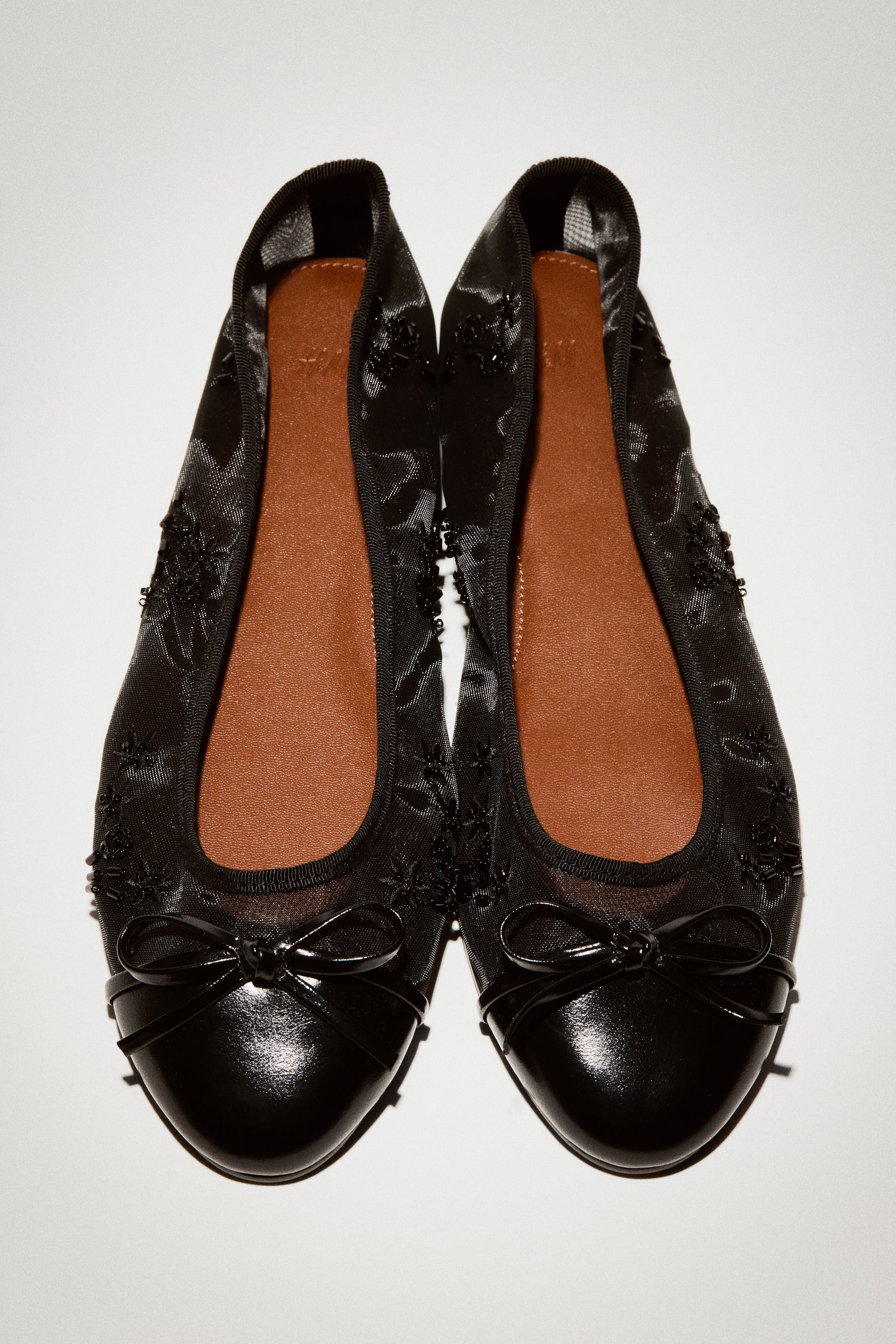 View larger image: A pair of black mesh ballet flats is shown from a top-down view, featuring semi-sheer uppers embellished with matching black beads. The shoes are displaying glossy, coated-fabric bows on solid black almond toe caps, finished with a black grosgrain trim and contrasting tan insoles.