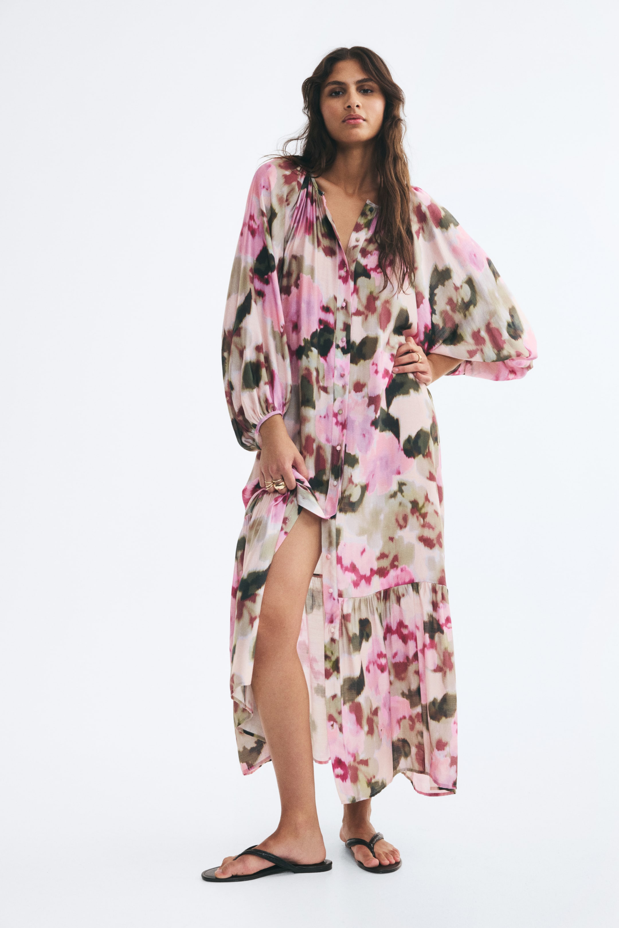 Ampliar la imagen: Una modelo lleva puesto un vestido túnica largo y oversize con un patrón abstracto en rosa y verde, mientras levanta el bajo hacia un lado. El vestido presenta mangas largas tipo globo, cierre frontal con botones y un panel fruncido por encima del dobladillo, y se combina con sandalias tipo thong negras.