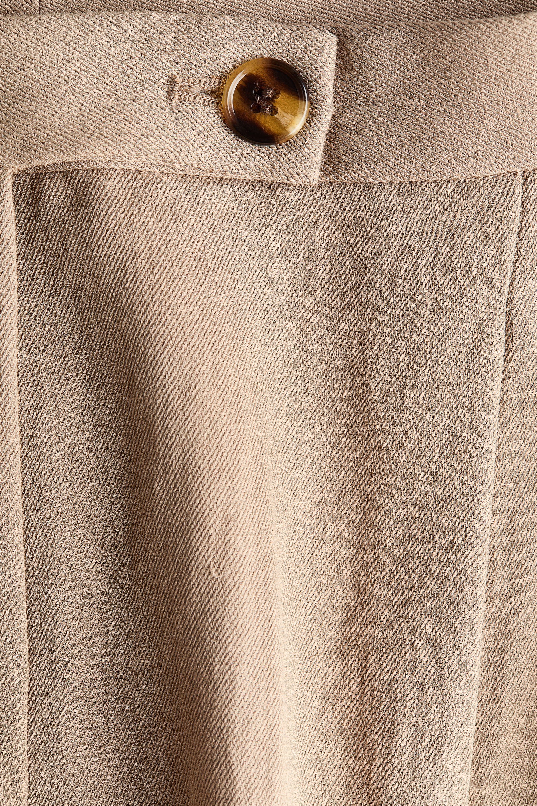 Se større billede: Beige vævede bukser har en forlænget linning med en brun skildpaddemønstret knap, og lodrette folder er synlige på stoffet nedenunder.