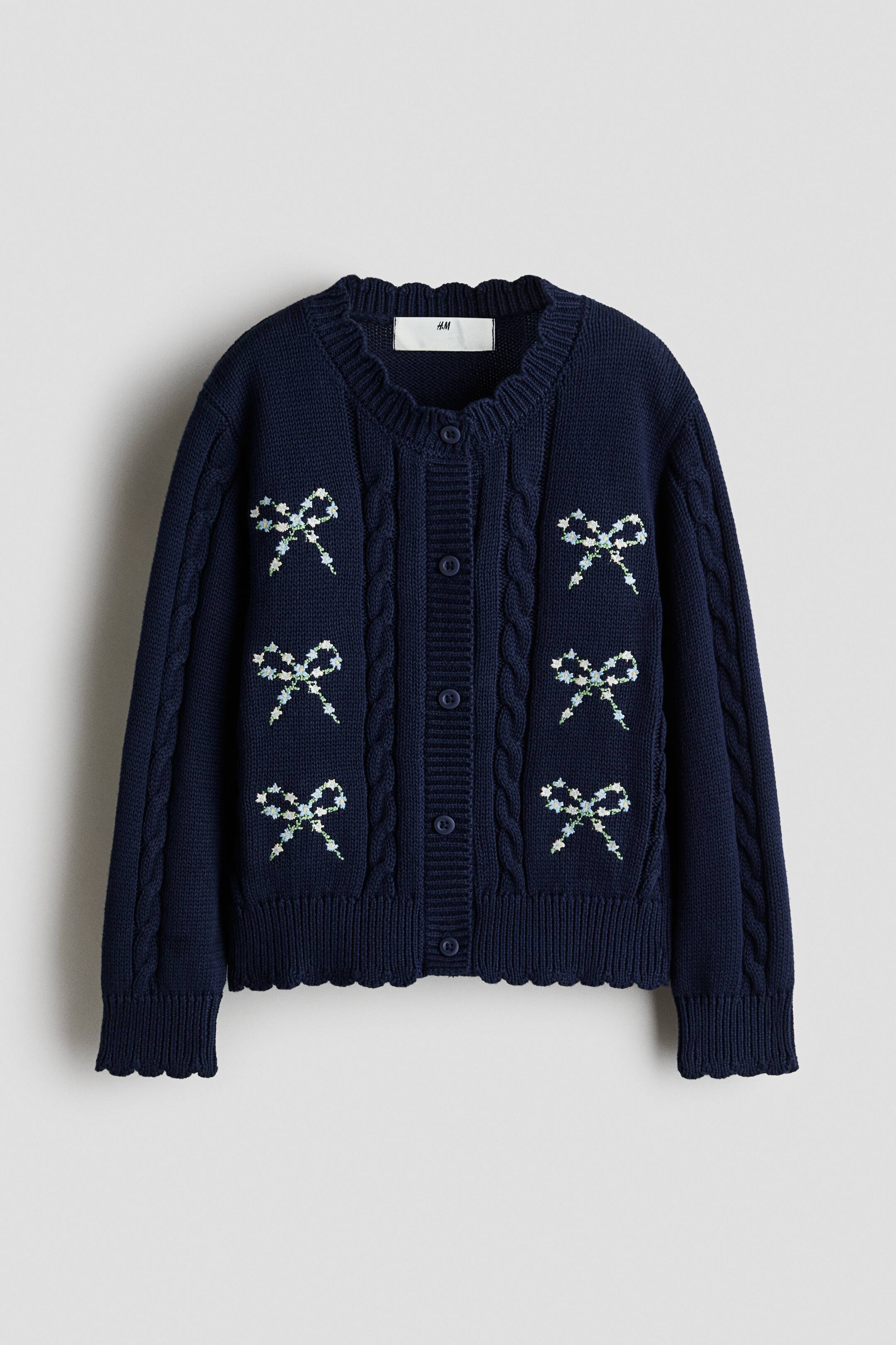 View larger image: A navy blue knit cardigan is shown from the front, featuring a round neckline, button-down closure, and cable-knit details along the front and sleeves. Six embroidered floral bow motifs decorate the chest area, with scalloped edges finishing the ribbed collar, cuffs, and hem.