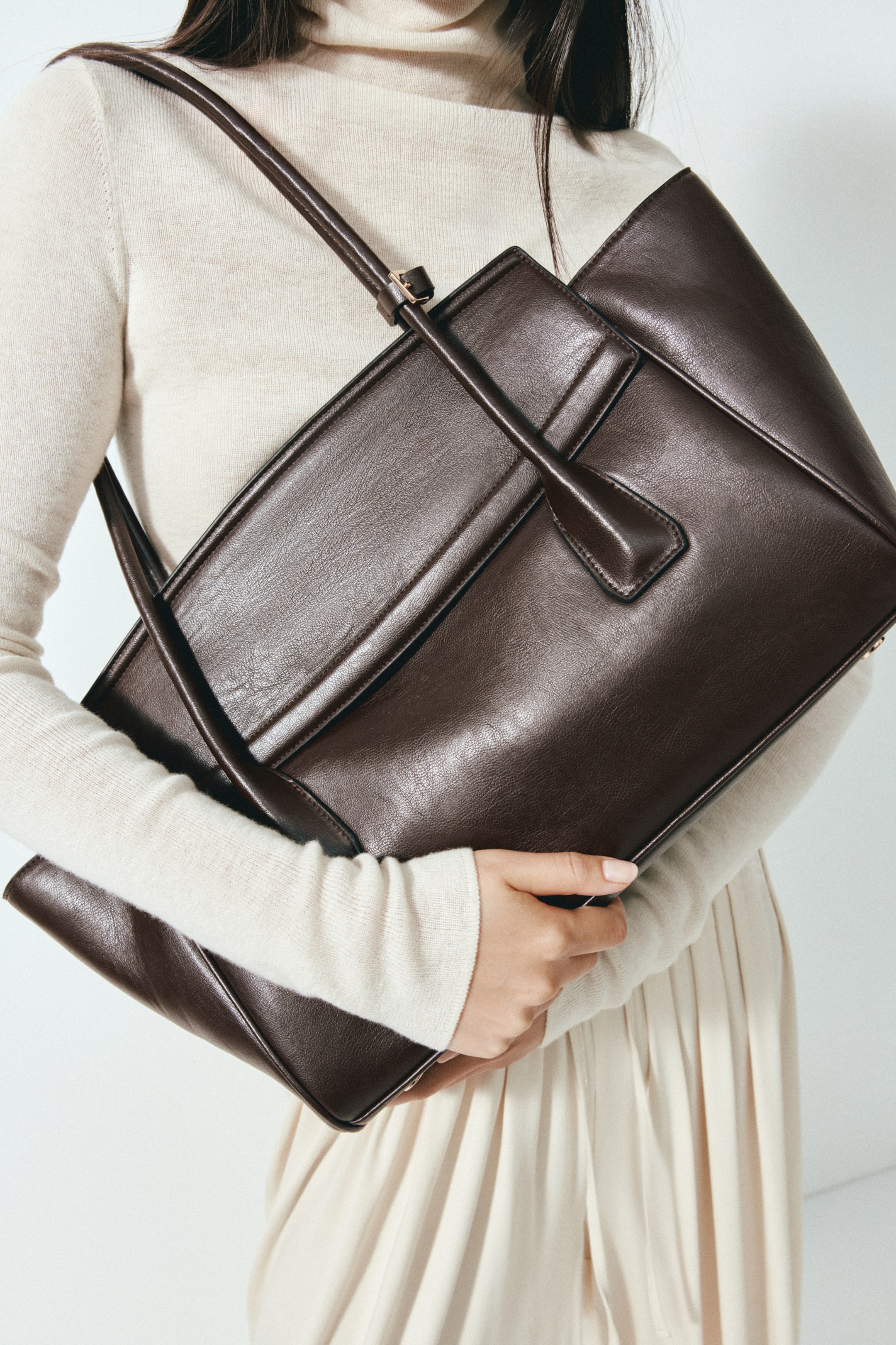 View larger image: A dark brown shoulder bag, with its long handles fastened together by a tab and a small metal buckle, is being carried by the model. One handle is draped over the model's shoulder, and the model's hand is holding the bag's bottom edge.