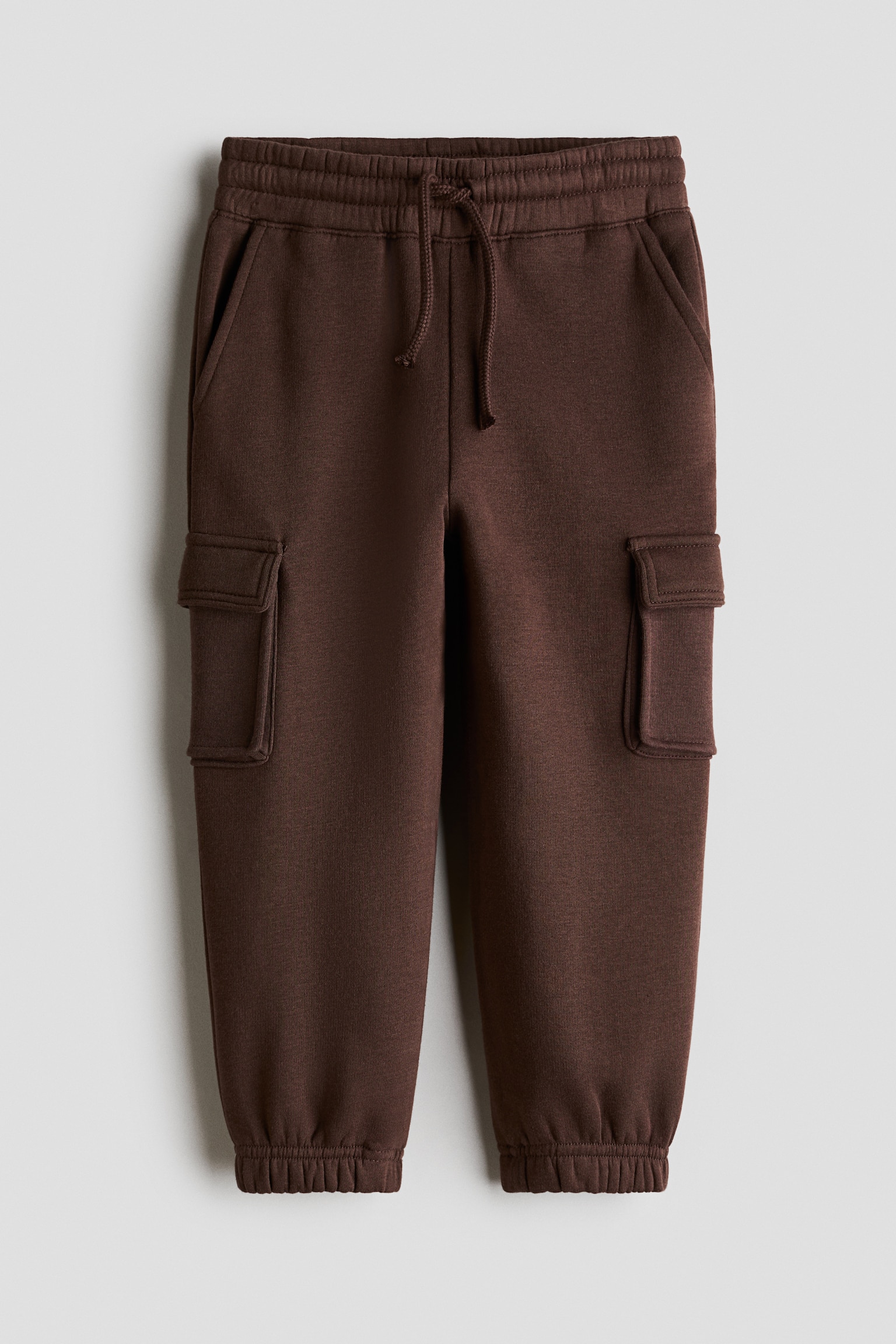 Un pantalon de jogging marron foncÃ© en molleton est prÃ©sentÃ© de face, avec une taille Ã©lastiquÃ©e munie dâun cordon de serrage et des poches latÃ©rales en biais. Le pantalon met en valeur des poches soufflet Ã  rabat sur les cuisses et se termine par des bas de jambe Ã©lastiquÃ©s aux chevilles.