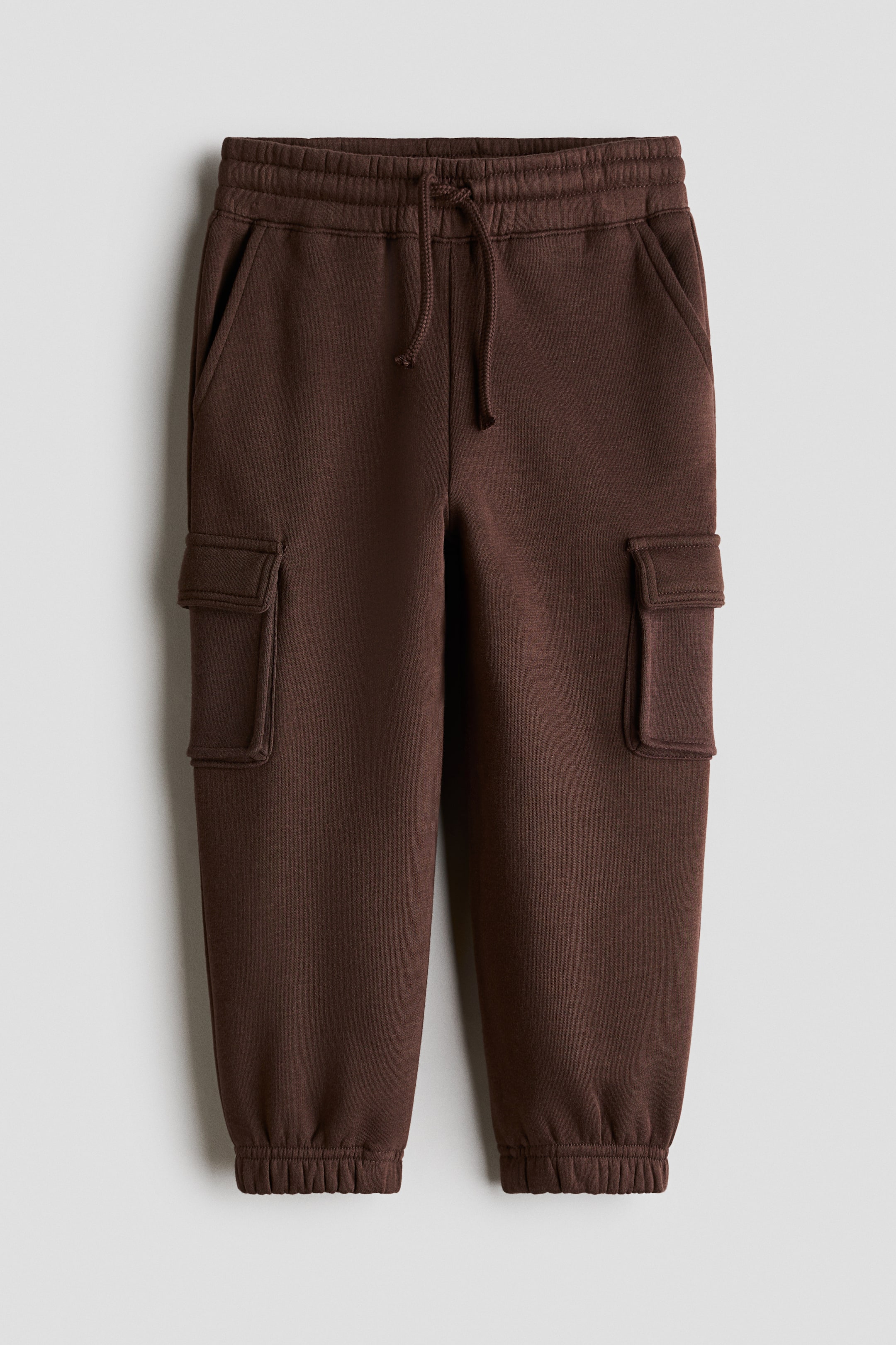 Größeres Bild anzeigen: Dunkelbraune Trainingshose aus Sweatstoff, von vorne dargestellt, mit elastischem Bund und Kordelzug sowie schrägen Seitentaschen. Die Hose zeigt aufgesetzte Beintaschen mit Patten an den Oberschenkeln und schließt an den Knöcheln mit elastischen Bündchen ab.