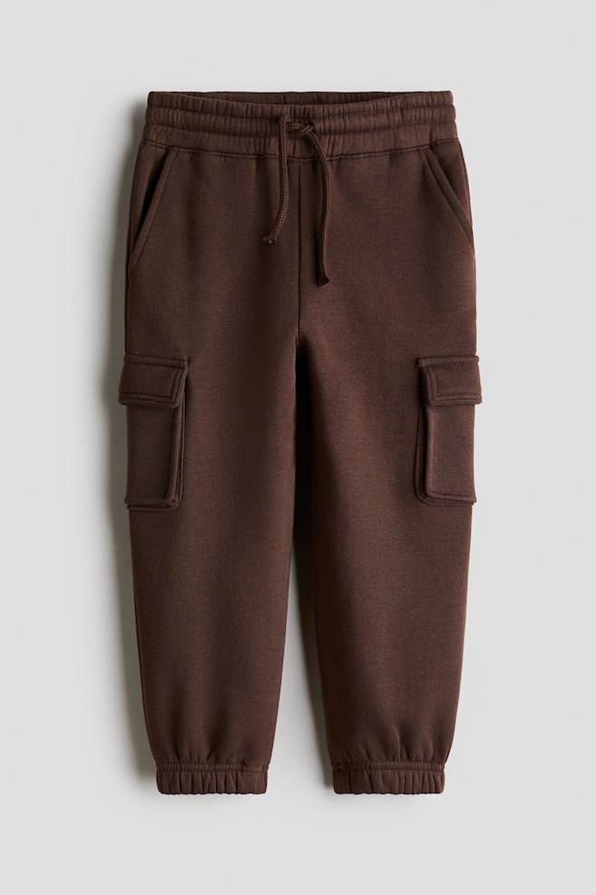 Dunkelbraune Jogginghose aus Sweatstoff, von vorne dargestellt, mit elastischem Bund und Kordelzug sowie schrÃ¤gen Seitentaschen. Die Hose zeigt aufgesetzte Blasebalgtaschen mit Klappen an den Oberschenkeln und schlieÃt an den KnÃ¶cheln mit elastischen BÃ¼ndchen ab. - 1