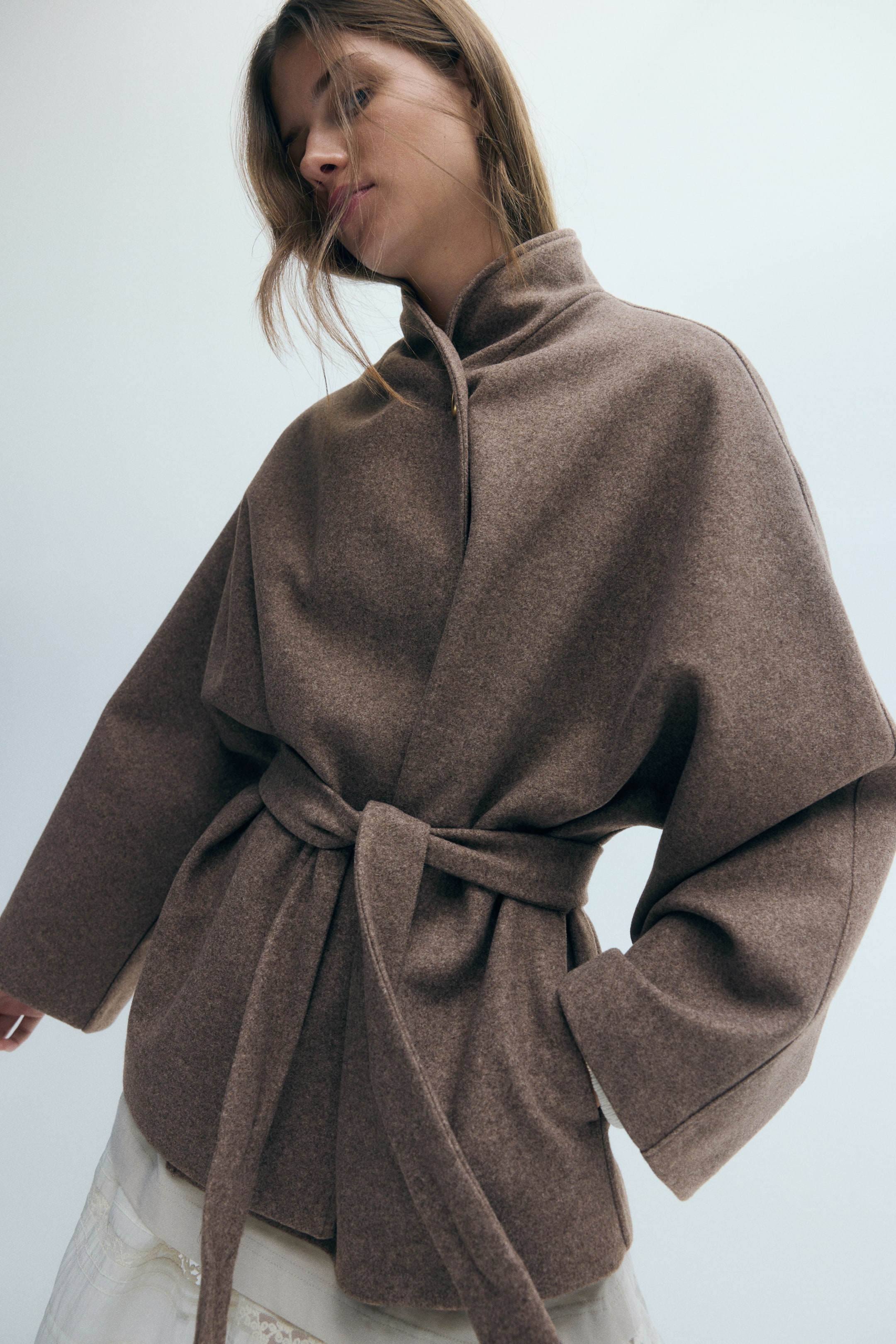 Zobraziť väčší obrázok: Model má na sebe oversized hnedý kabát z textúrovanej plstenej látky, s očkom stojatým golierom, širokými rukávmi a opaskom v rovnakom odtieni uviazaným v páse. Jednu ruku má založenú vo vrecku na boku a spod lemovania je viditeľná svetlá sukňa.