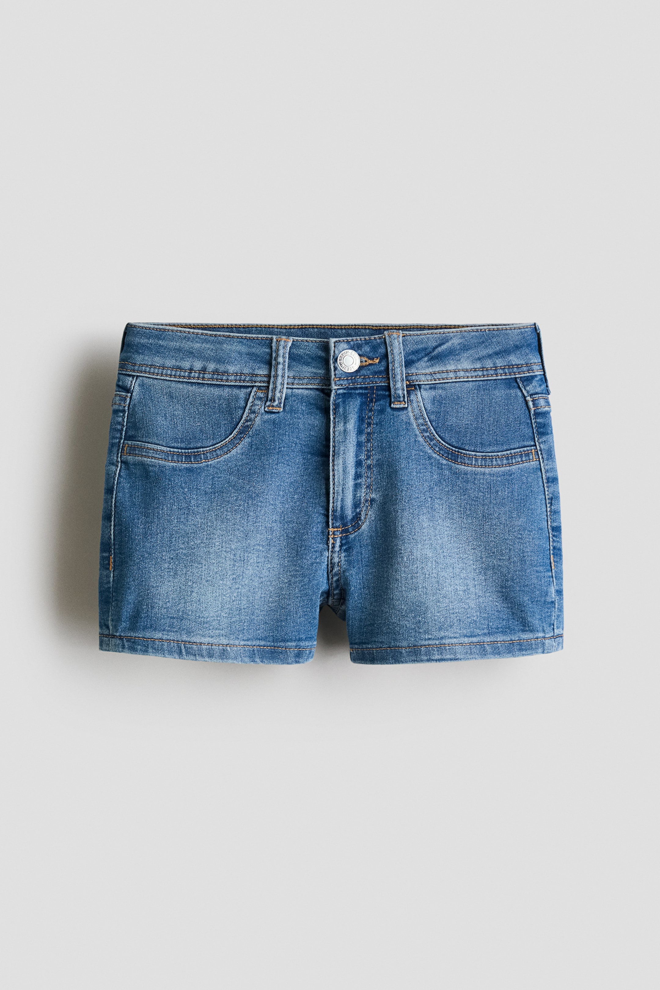 Grösseres Bild ansehen: Ein Paar mittelblaue Jeansshorts ist von vorne zu sehen, mit einem flachen Bund, der fünf Gürtelschlaufen aufweist, sowie einem silberfarbenen Knopf und Reißverschluss. Unter dem Bund sind zwei geschwungene Fronttaschen sichtbar, und die Shorts zeigen gerade gestickte Säume.