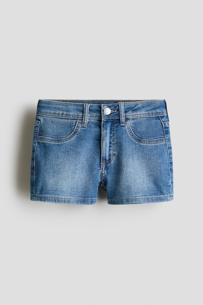 Ein Paar mittelblaue Jeansshorts ist von vorne zu sehen, mit einem flachen Bund, der fünf Gürtelschlaufen aufweist, sowie einem silberfarbenen Knopf und Reißverschluss. Unter dem Bund sind zwei geschwungene Fronttaschen sichtbar, und die Shorts zeigen gerade gestickte Säume. - 1