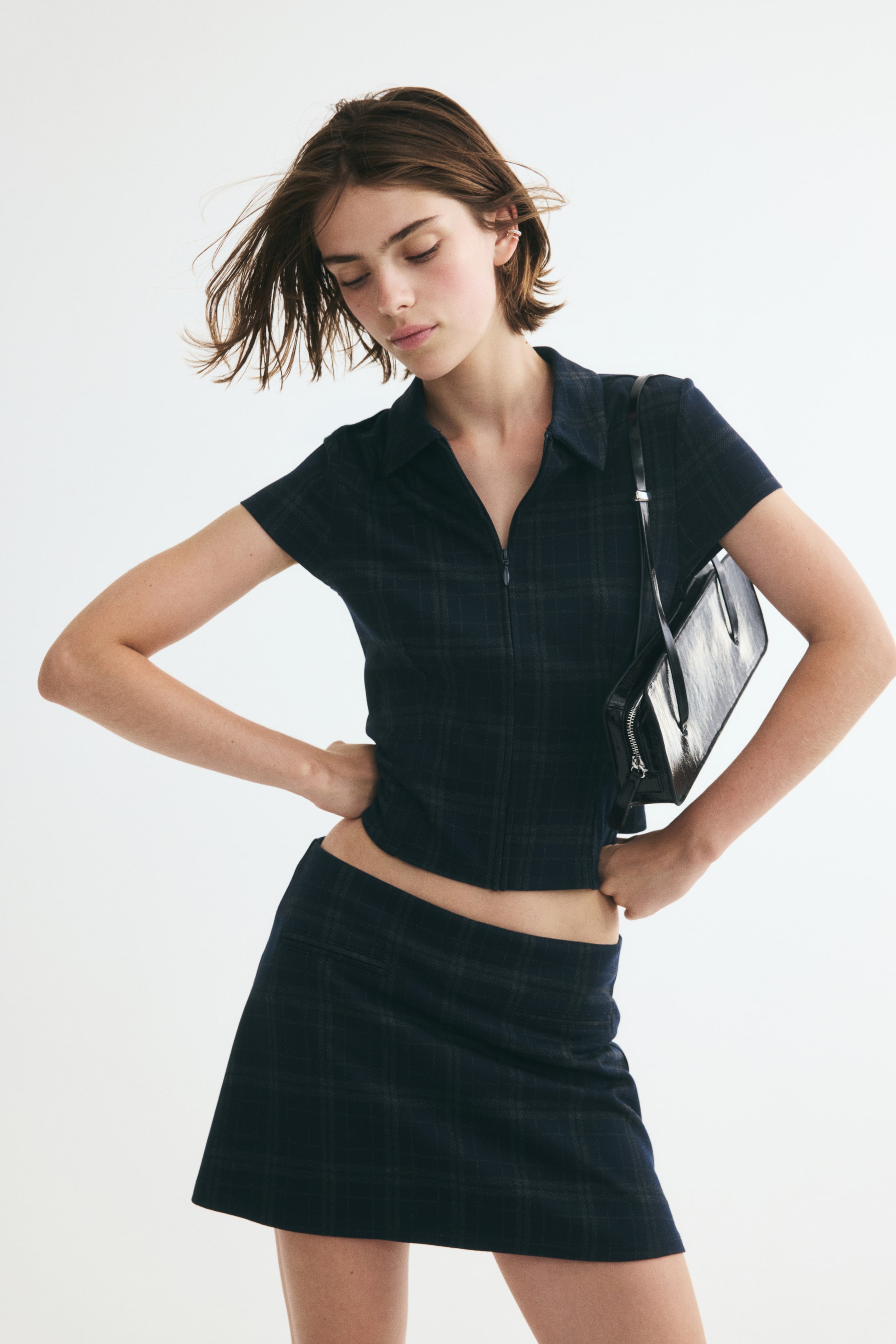 Ampliar la imagen: La modelo lleva puesta una blusa oscura con estampado de cuadros, cuello, mangas cortas tipo cap y cierre frontal con cierre de cremallera. La blusa se combina con una minifalda a juego de cuadros, y lleva un bolso negro al hombro.