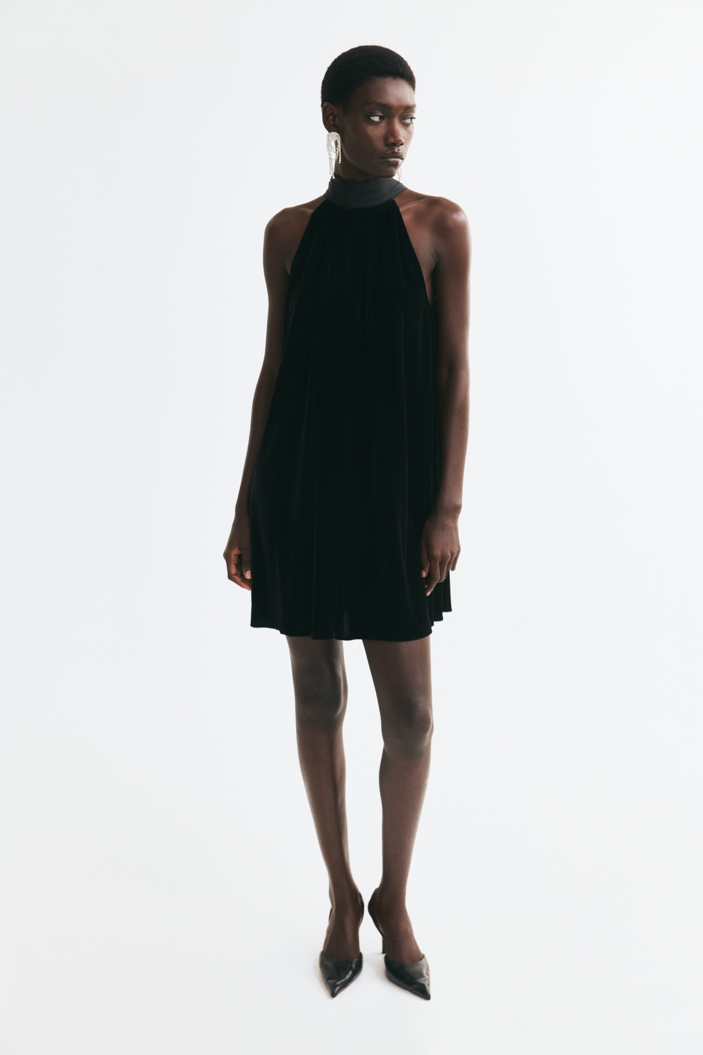 A black velvet halter dress is hanging in a front-facing view, featuring a high, gathered satin collar and deep-cut sleeveless armholes. The dress drapes downward in a loose A-line silhouette with subtle pleating starting from the neckline.