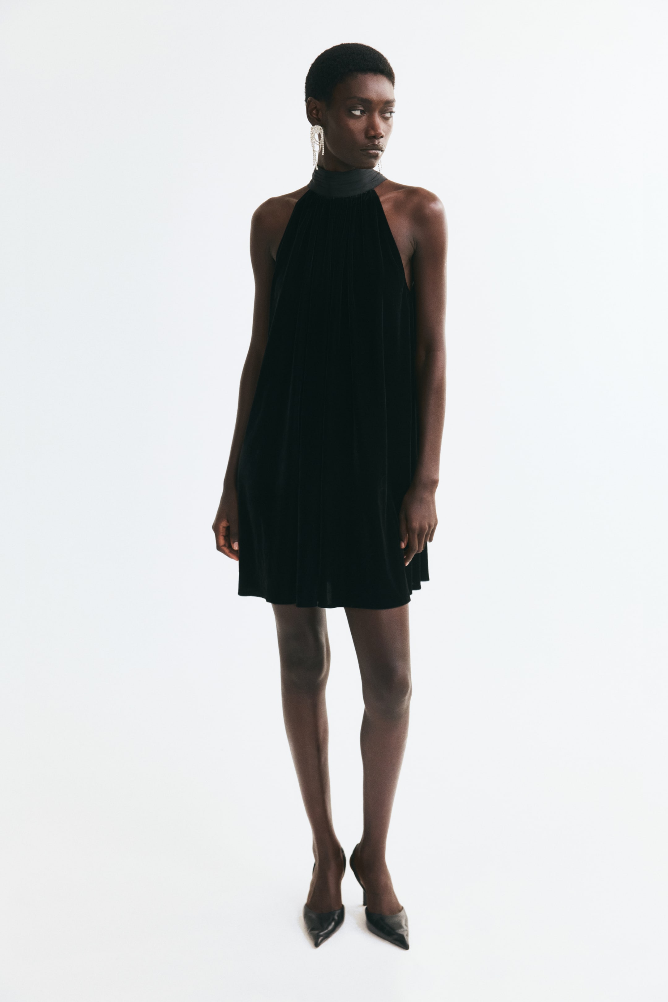View larger image: A short black A-line dress with a pleated high stand collar and a sleeveless halter-neck cut is worn by the model. The loose-fit dress is styled with black pointed-toe heels.