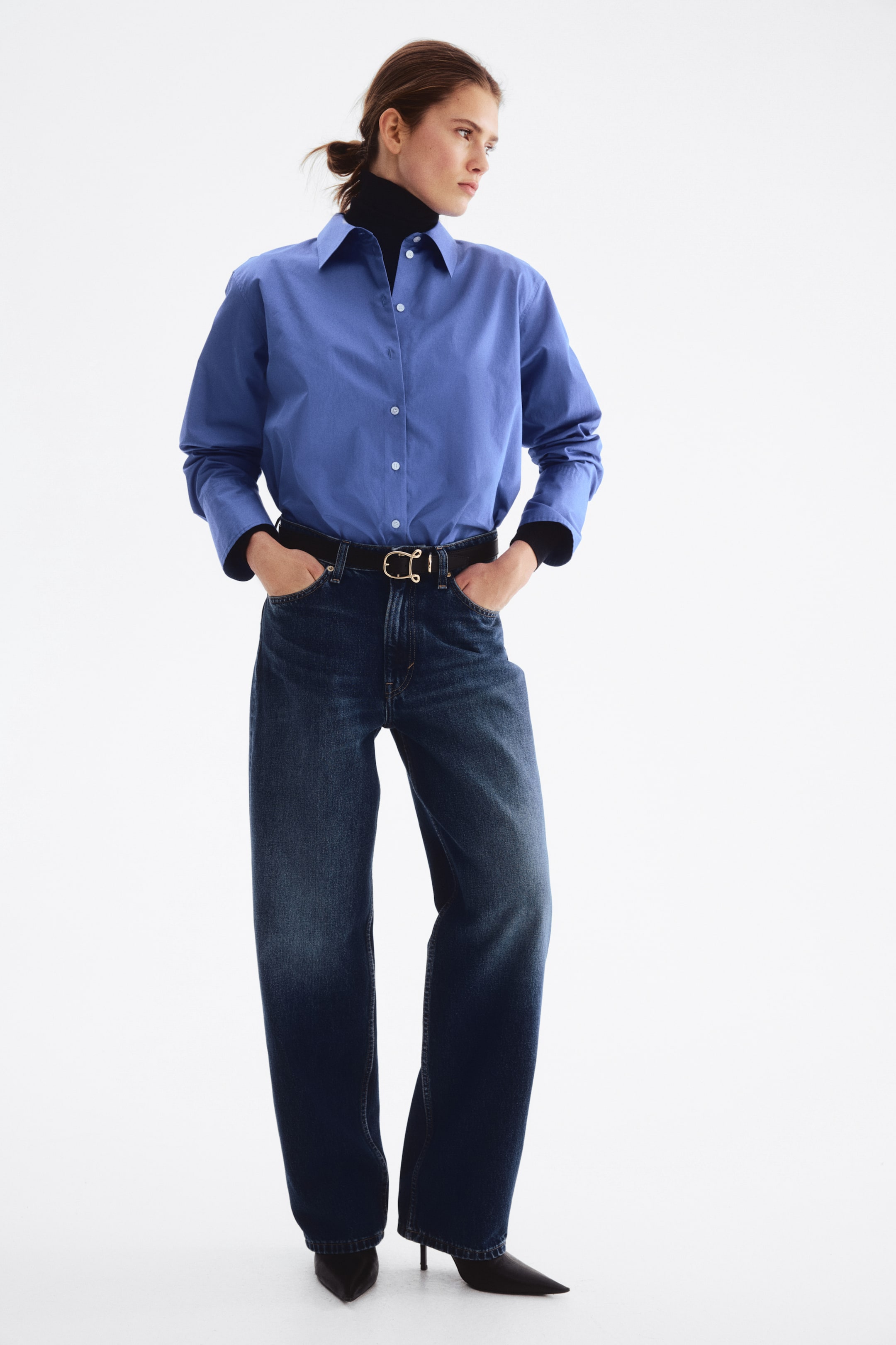 Ver imagen más grande: Una camisa azul de botones se superpone a un jersey de cuello alto negro, presentando un ajuste relajado y un cuello clásico. Se llevan unos vaqueros de pierna ancha y lavado oscuro, ajustados en la cintura con un cinturón negro con hebilla dorada, y unos zapatos de tacón con punta negra completan el look.