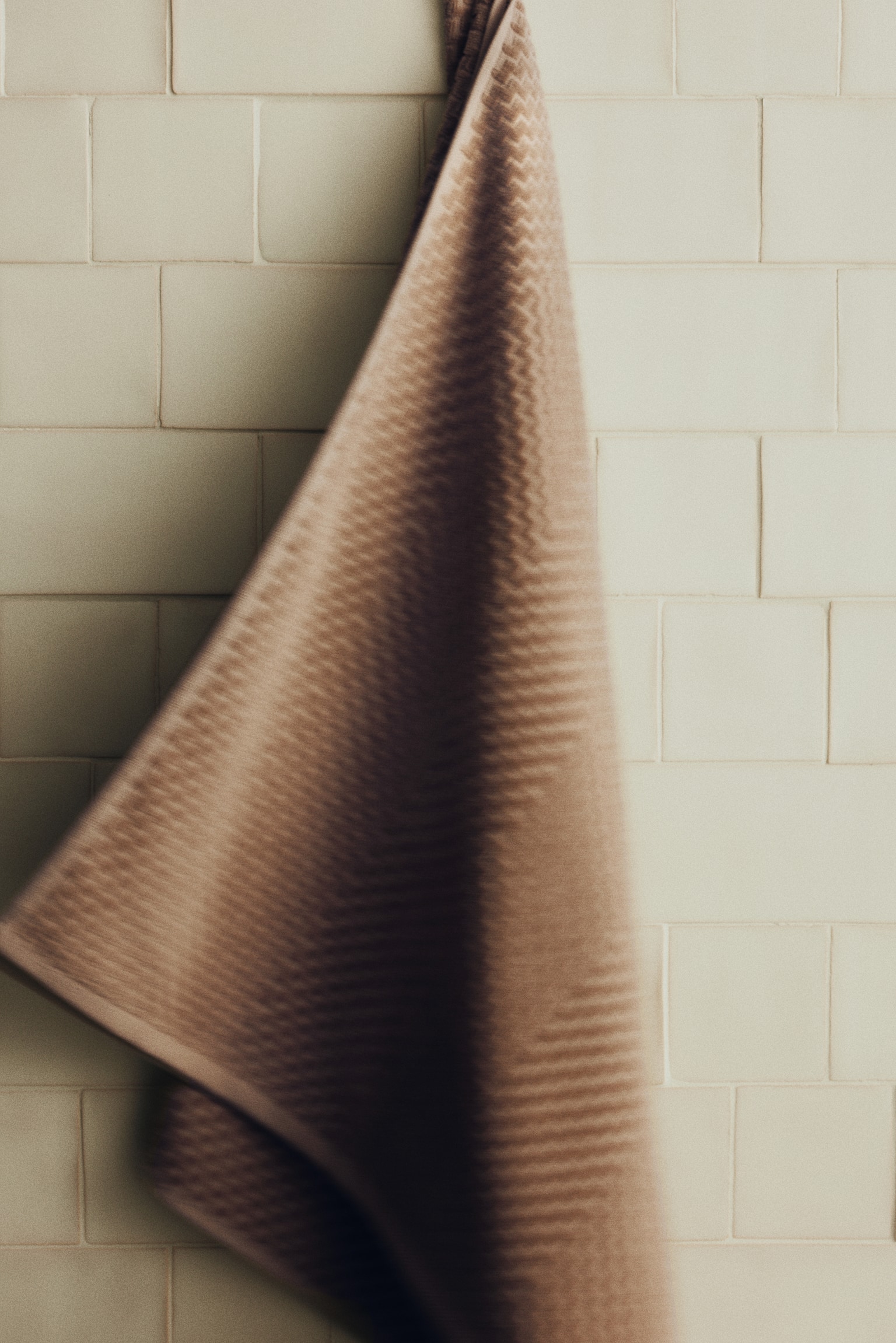 Une serviette de bain marron clair, arborant un motif en zigzag texturé, est suspendue à un crochet sombre par sa boucle intégrée. La serviette est pliée, révélant ses couches et sa bordure subtile. - 1