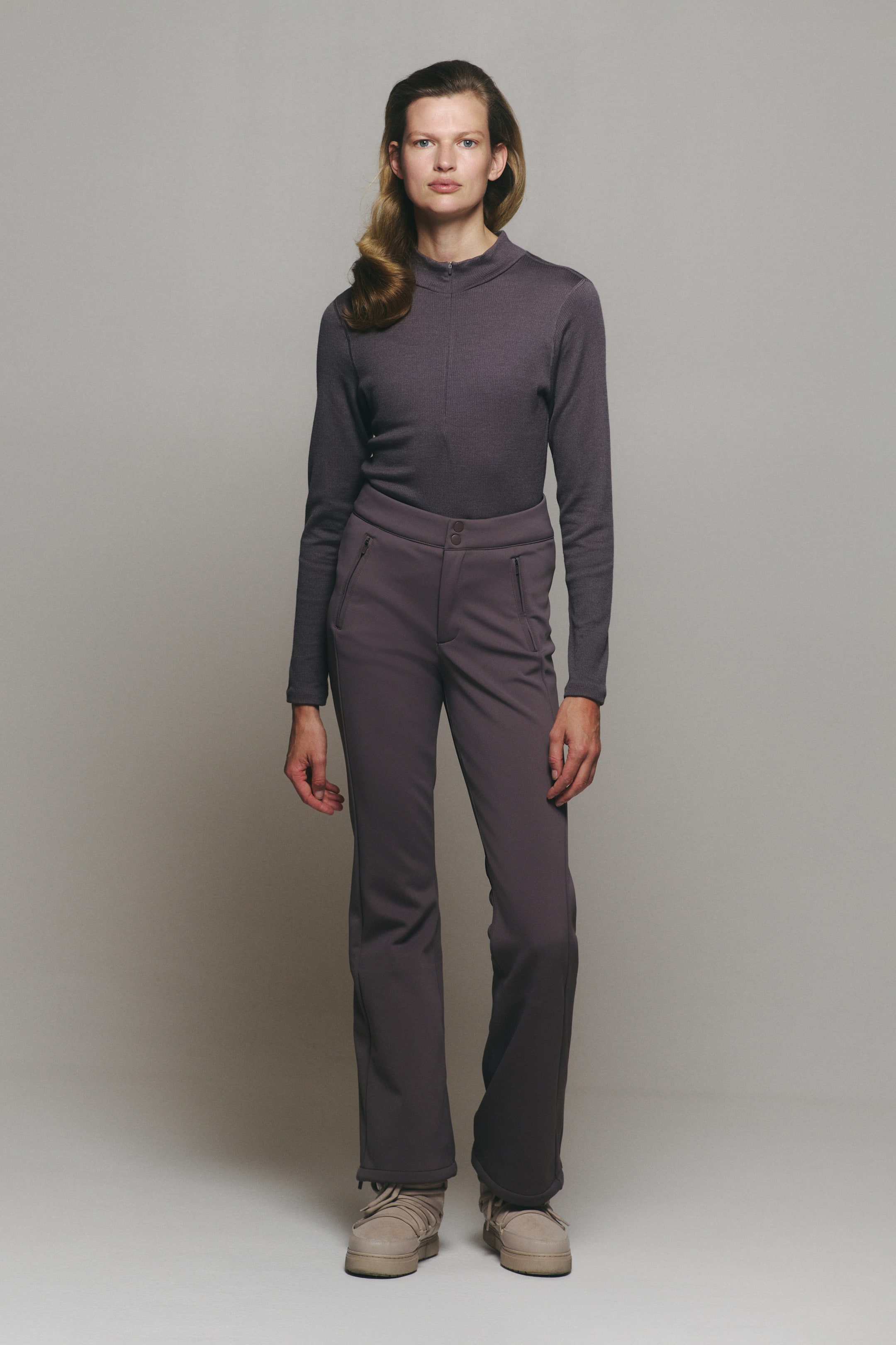 View larger image: A dark purple outfit consisting of a ribbed, long-sleeved mock-neck top with a front zipper and matching slim-fit trousers is being worn by the model. The top is tucked into the high-waisted trousers, which feature side zipper pockets and a double-button closure.