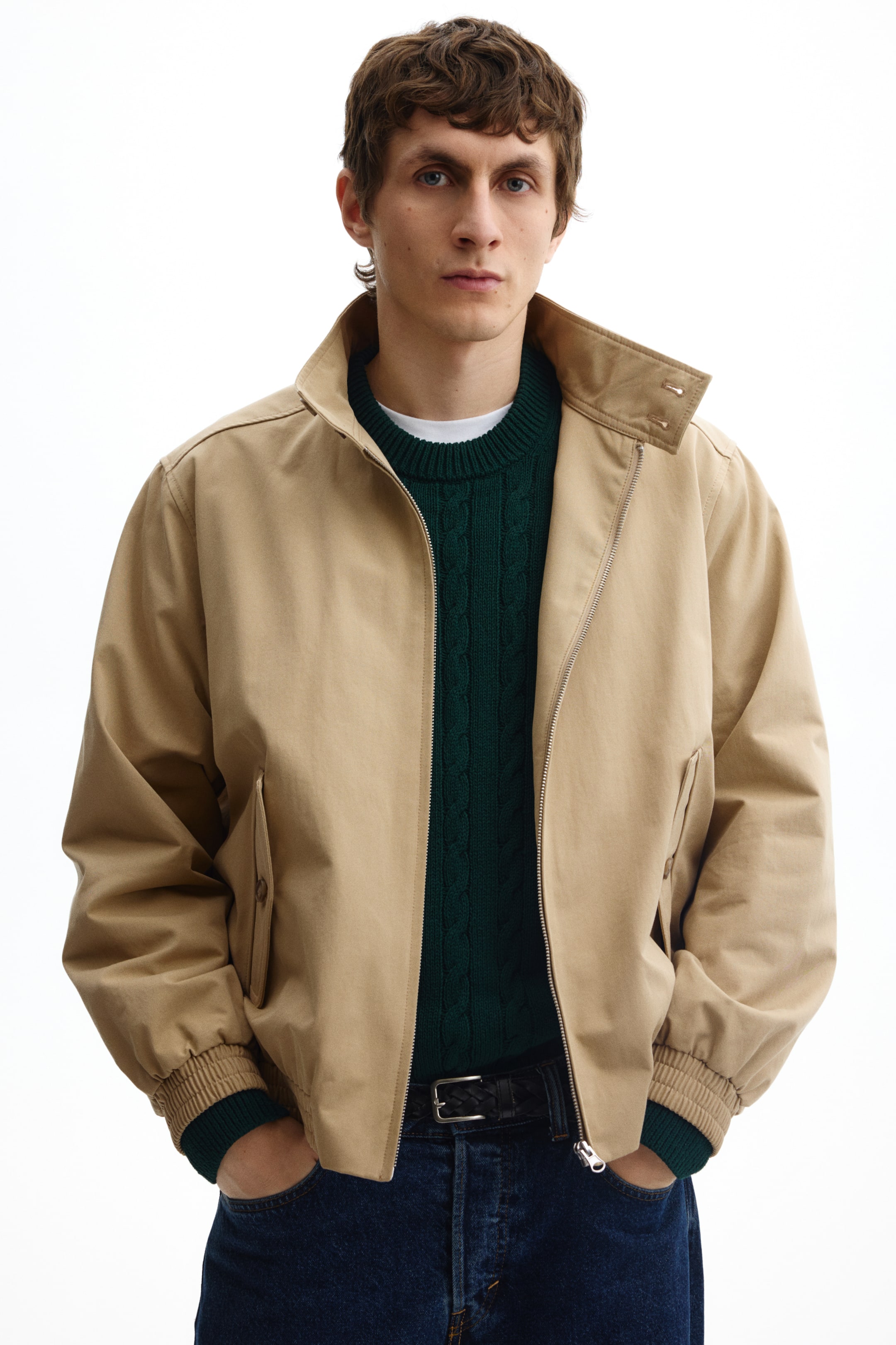 Se større billede: En lysebrun bomberjakke præsenteres med en stående krave med to trykknapper, en sølvfarvet lynlås i fronten samt elastiske manchetter og kant. Jakken bæres åben og afslører en mørkegrøn kabelstrikket sweater nedenunder.