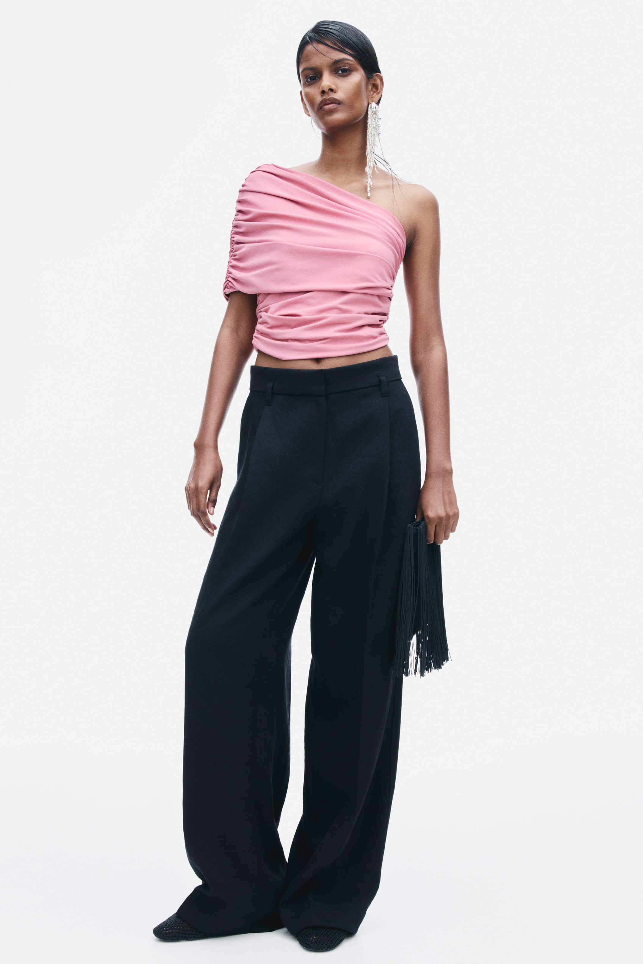 View larger image: A pink, one-shoulder cropped top featuring draped and gathered detailing is being worn by the model. The top is styled with high-waisted, wide-leg black trousers, and the model is holding a black fringed clutch.