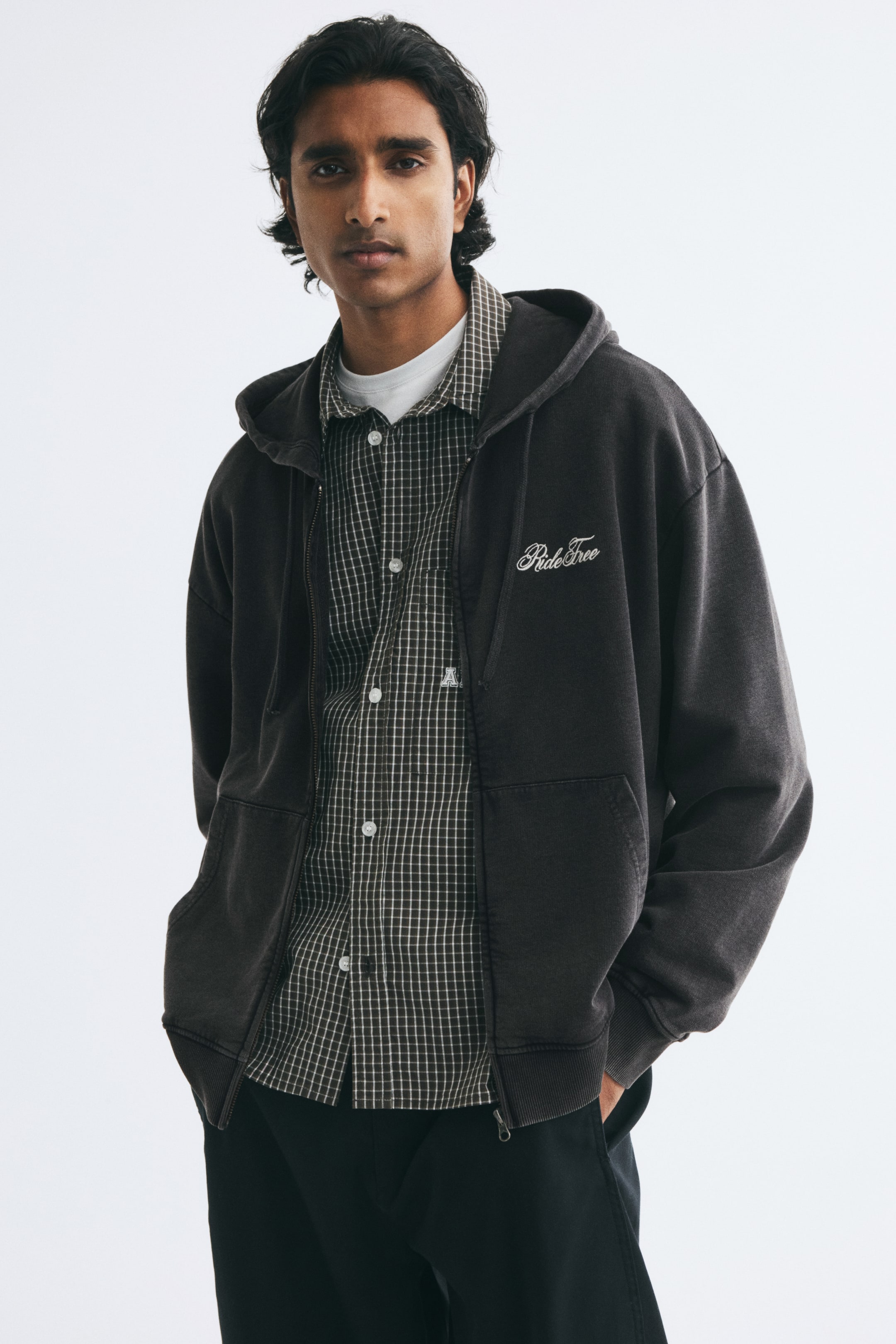 View larger image: A faded black zip-up hoodie featuring white cursive embroidery on the chest is being worn open by the model. The hoodie is styled over a dark plaid button-up shirt and a white T-shirt, paired with black trousers.