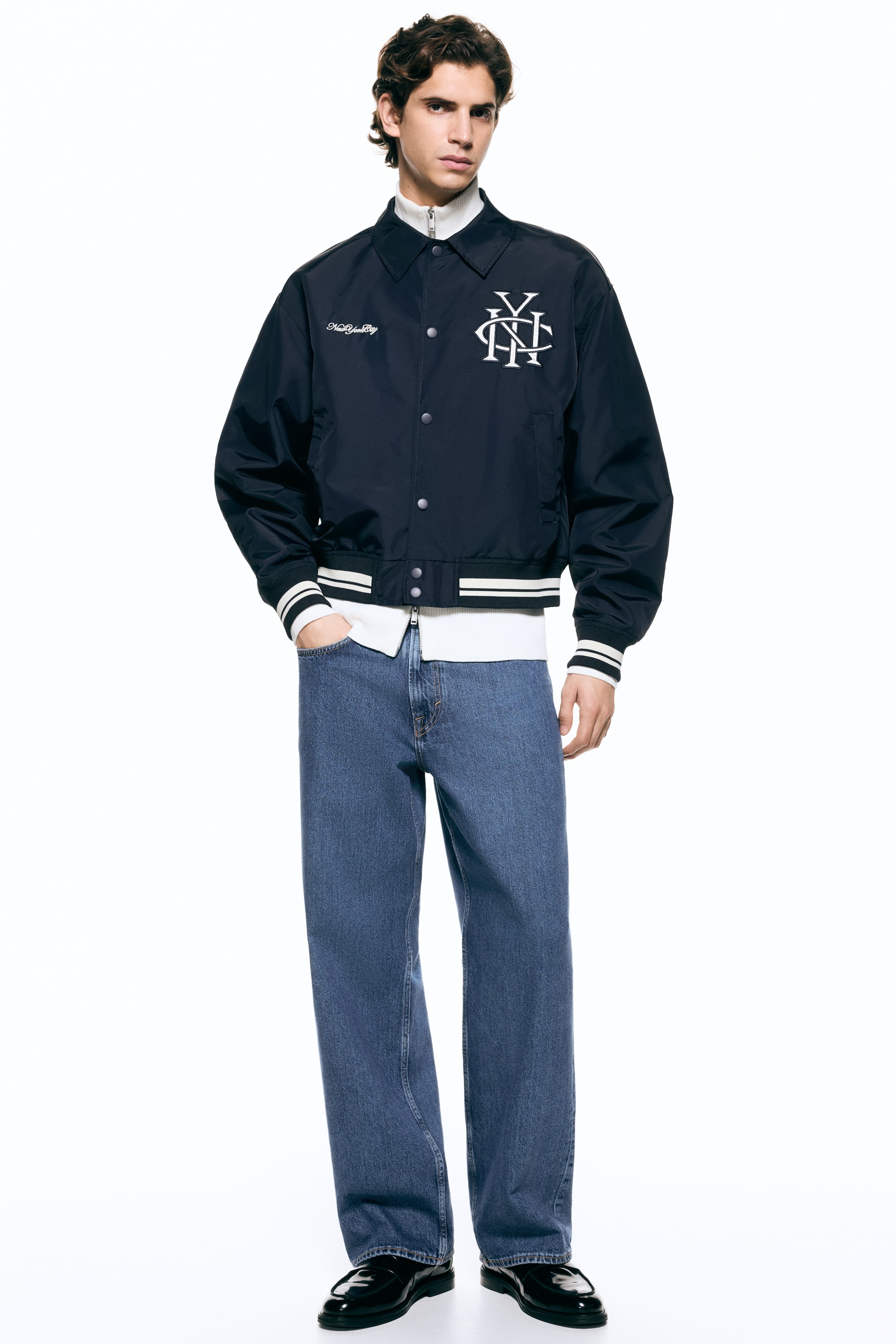 View larger image: A dark blue bomber jacket is facing forward, featuring a pointed collar, silver snap buttons down the front, and two angled welt pockets. It is showcasing ribbed cuffs and a matching ribbed hem, both detailed with two white stripes, and displaying white embroidered "New York City" text on the left chest and a large white embroidered interlocking "NY" monogram on the right chest, while a white ribbed mock-neck top is visible underneath.