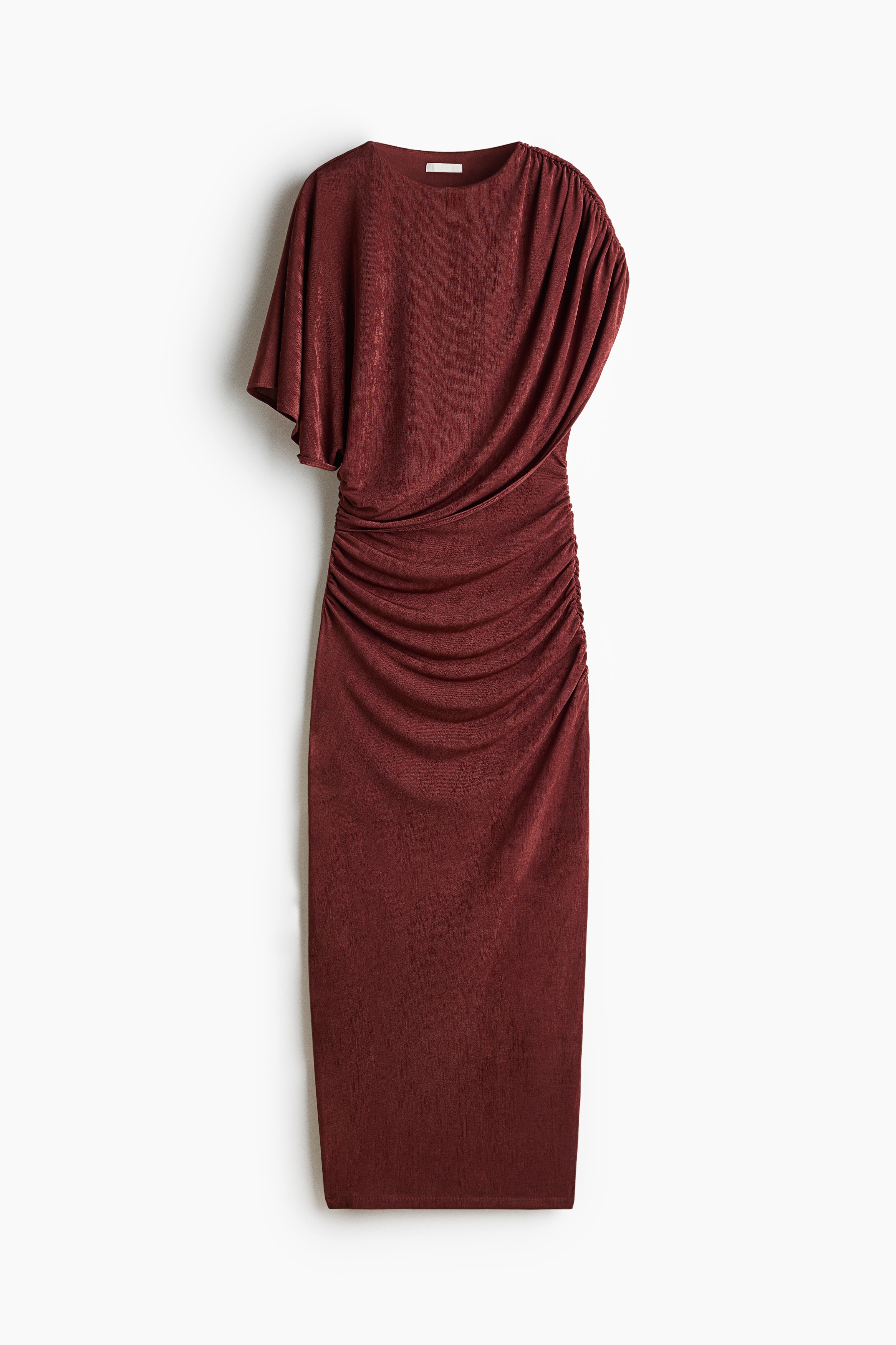 A rust-colored, fitted dress in stretch jersey is being worn by the model, featuring a round neckline and an asymmetrical design. The dress has a draped, cape-like layer on one shoulder and gathered, ruched seams along the bodice and hips to create a fitted, textured look.