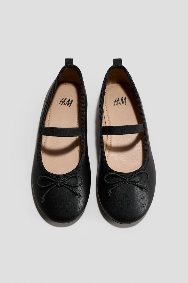 Ein Paar schwarze Ballerina-Pumps zeichnet sich durch abgerundete Zehen, einen elastischen Riemen über dem Spann und eine dekorative Schleife an der Vorderseite jeder Zehe aus. Die hellen Innensohlen zeigen ein sichtbares H&M-Logo. - 1