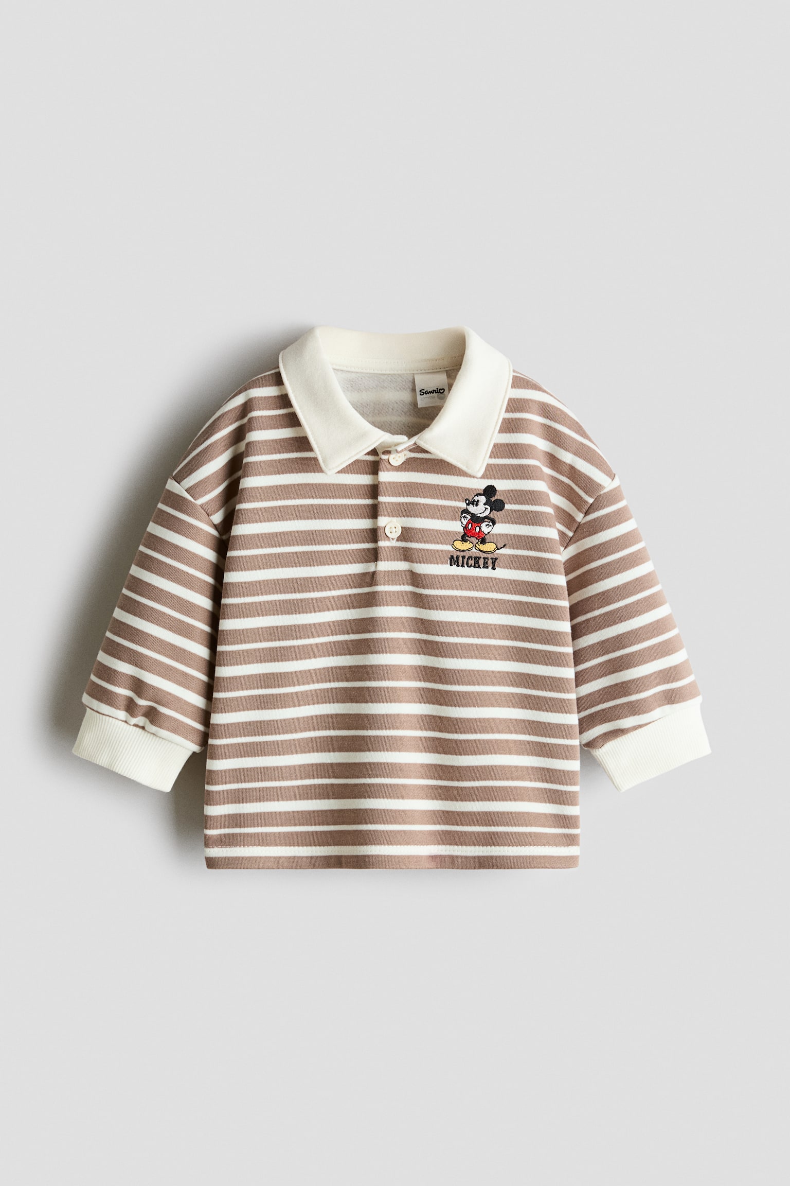 Una felpa polo a maniche lunghe, vista frontalmente, presenta righe orizzontali marrone chiaro e bianche, un colletto bianco tinta unita e polsini a coste bianchi coordinati. Mostra una chiusura con due bottoni e un ricamo di Mickey Mouse sul lato sinistro del petto con il nome "MICKEY" cucito sotto.