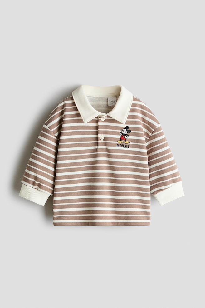 A striped rugby shirt is shown from the front, featuring a beige and white horizontal stripe pattern with a solid cream-coloured collar and matching wide ribbed cuffs. The shirt features a two-button placket, dropped shoulders, and a small embroidered Mickey Mouse detail on the left chest. - 1