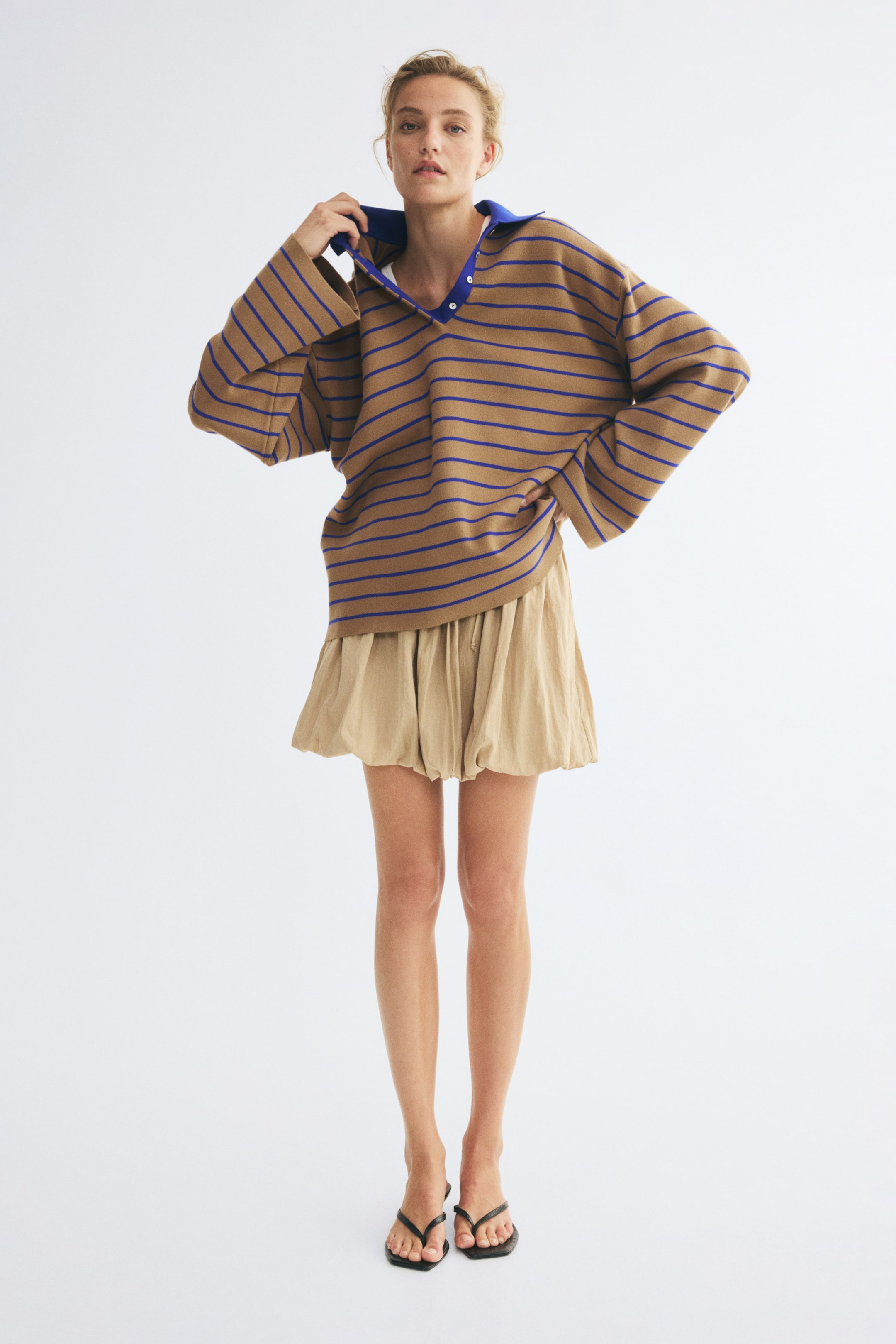 View larger image: An oversized polo shirt is shown from the front, featuring a relaxed, slouchy fit in a brown knit fabric with thin horizontal blue stripes. The shirt features a contrasting solid blue collar with a partially open button placket and wide, elongated sleeves that drape loosely.