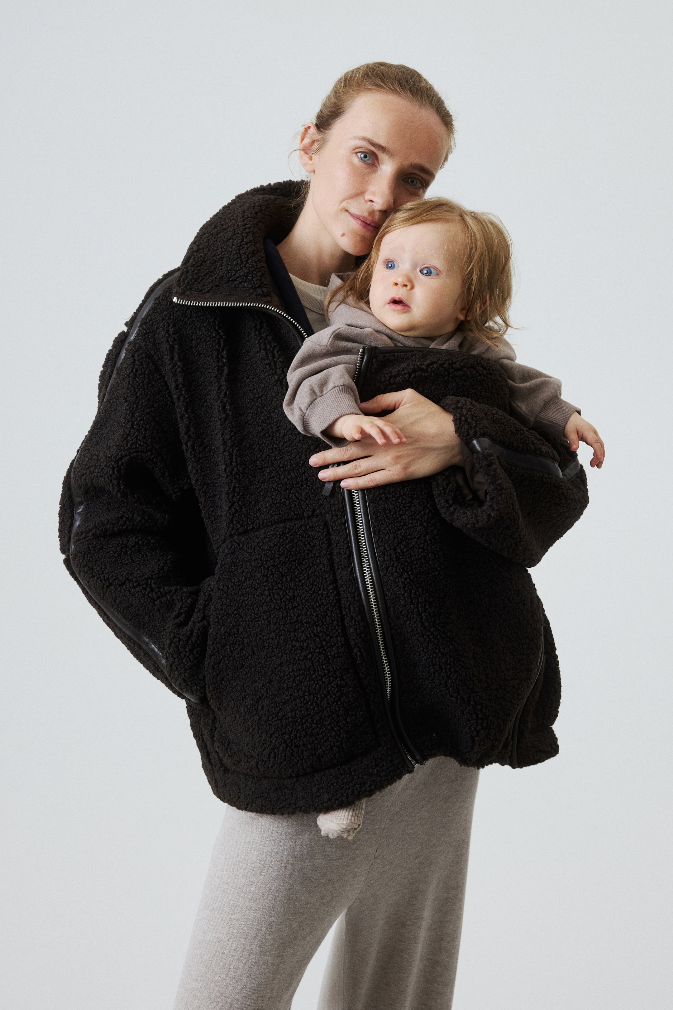 Se større billede: En sort oversized teddy-fleecejakke bæres af modellen og har lynlåslukning foran samt mørke kontrastkanter. Modellen holder en baby, som er delvist omsluttet af jakken.