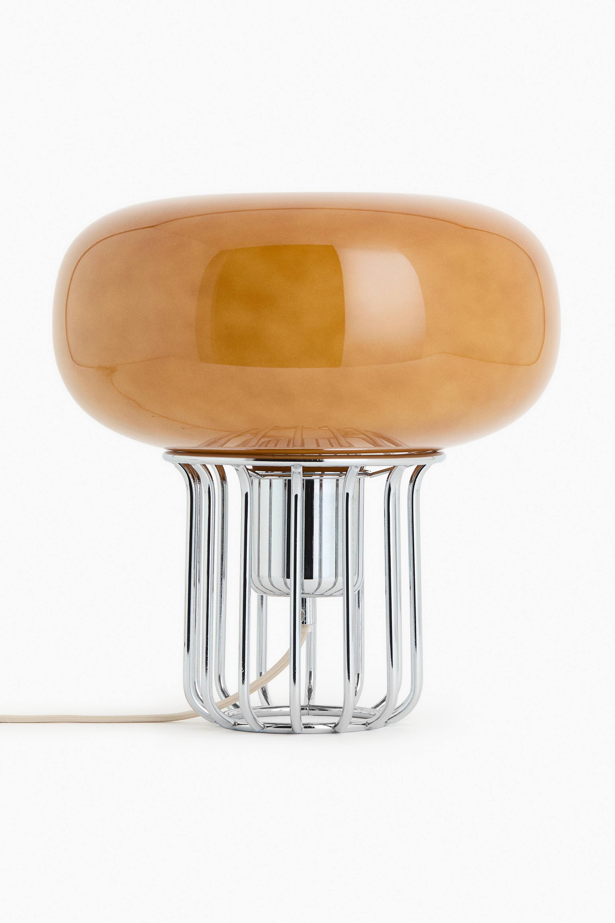 Größeres Bild ansehen: Glas-Tischlampe mit einem länglichen, bernsteinfarbenen Glasschirm, der auf einem verchromten Sockel ruht. Der Sockel bildet eine käfigartige Struktur mit vertikalen Metallstäben, und ein helles Stromkabel erstreckt sich von seinem Boden.