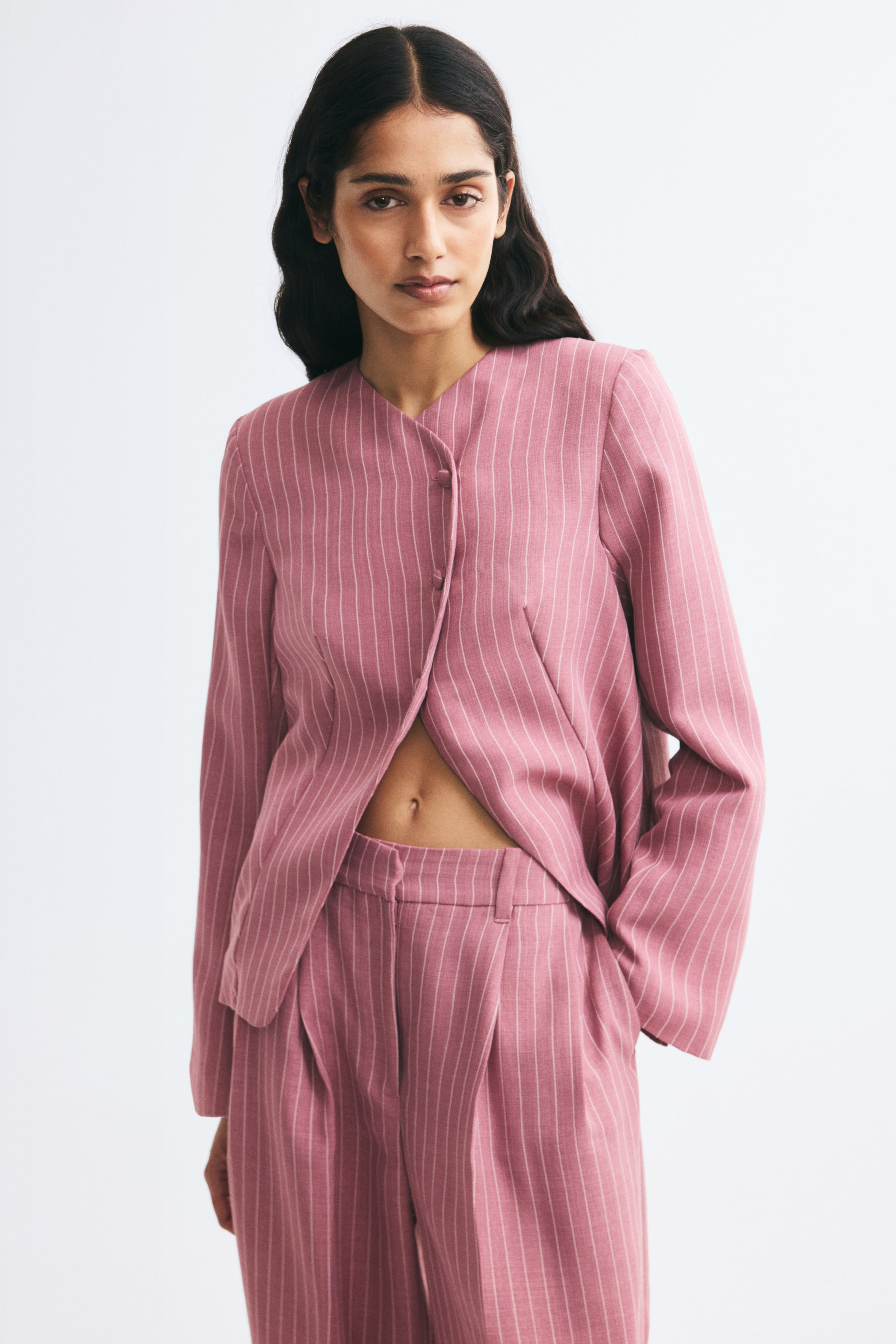 View larger image: A pink pinstripe single-breasted blazer is shown from the front, featuring a lapelless neckline and a slightly offset, angled front opening with covered buttons at the top. The blazer showcases structured shoulder pads and waist darts, parting at the waist to reveal matching trousers.
