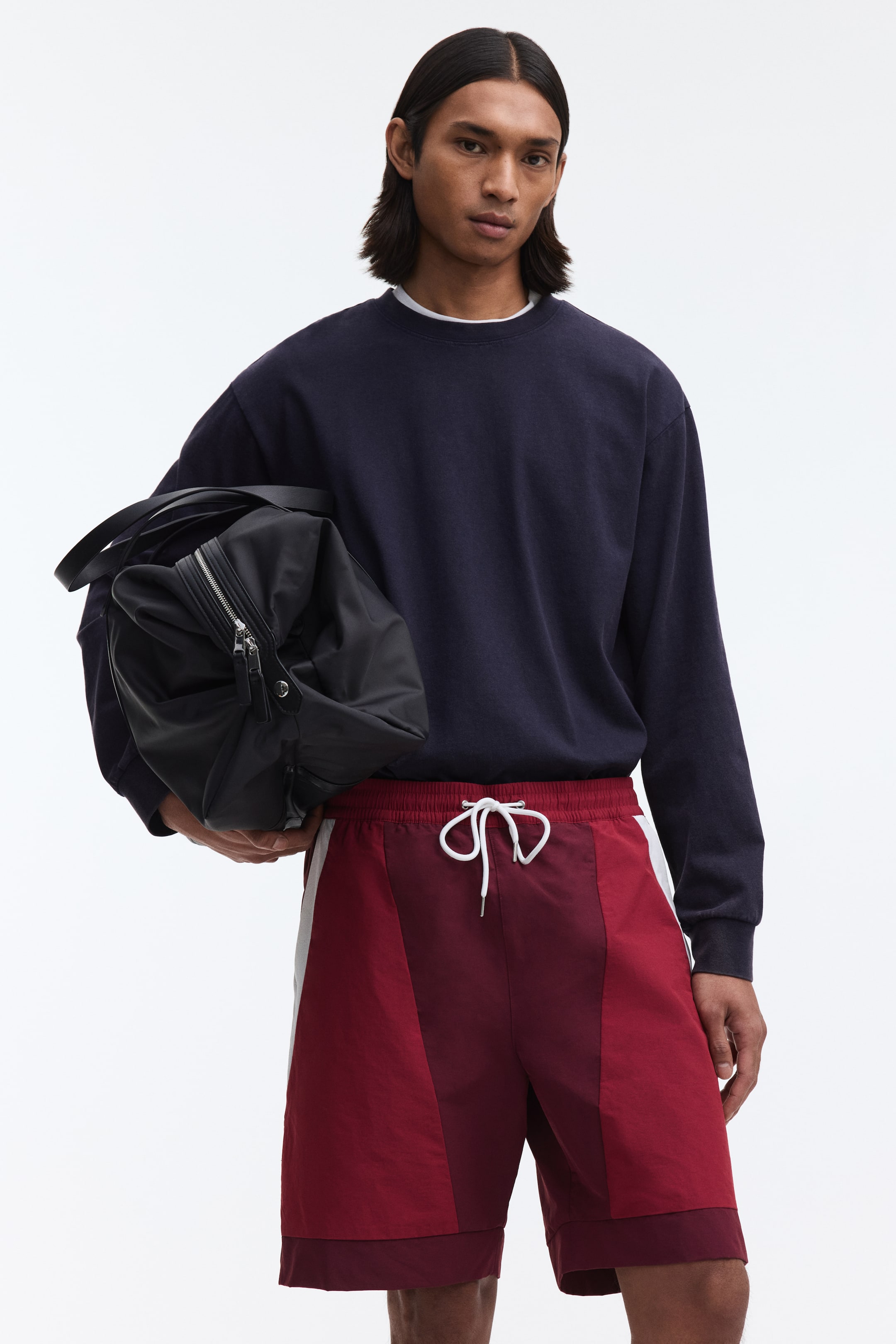 View larger image: The model is wearing track shorts with a relaxed fit, showcasing a two-toned burgundy design, accented with white side stripes and a white drawstring at the elasticated waist. A darker inner layer peeks out from beneath the hem, and the model is carrying a black duffel bag.