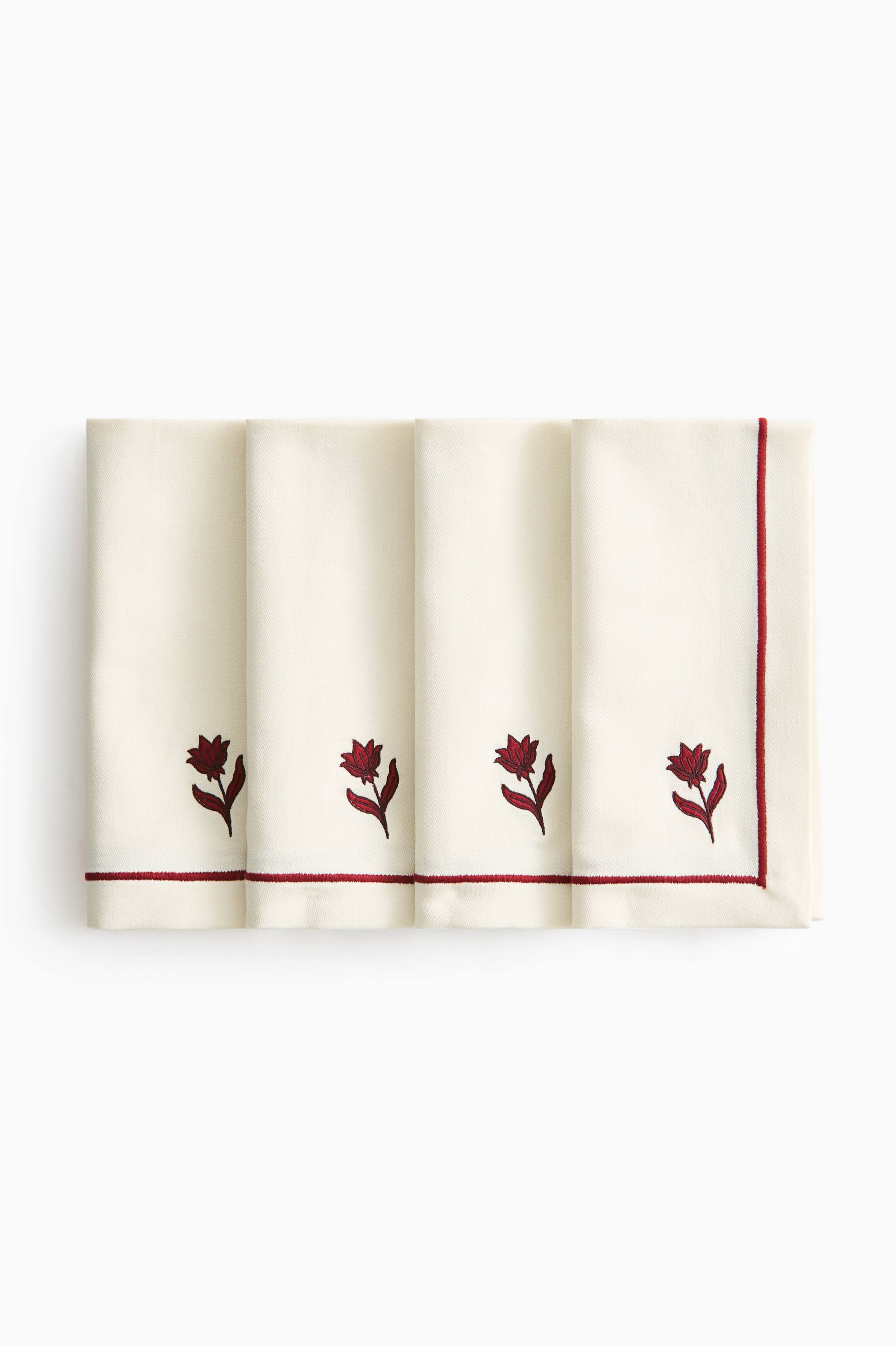 View larger image: A set of four cream-coloured cloth napkins is arranged in a flat-lay, overlapping row facing front. Each folded napkin shows a small, embroidered red tulip near the bottom, accented by a thin red embroidered border running along the hem.