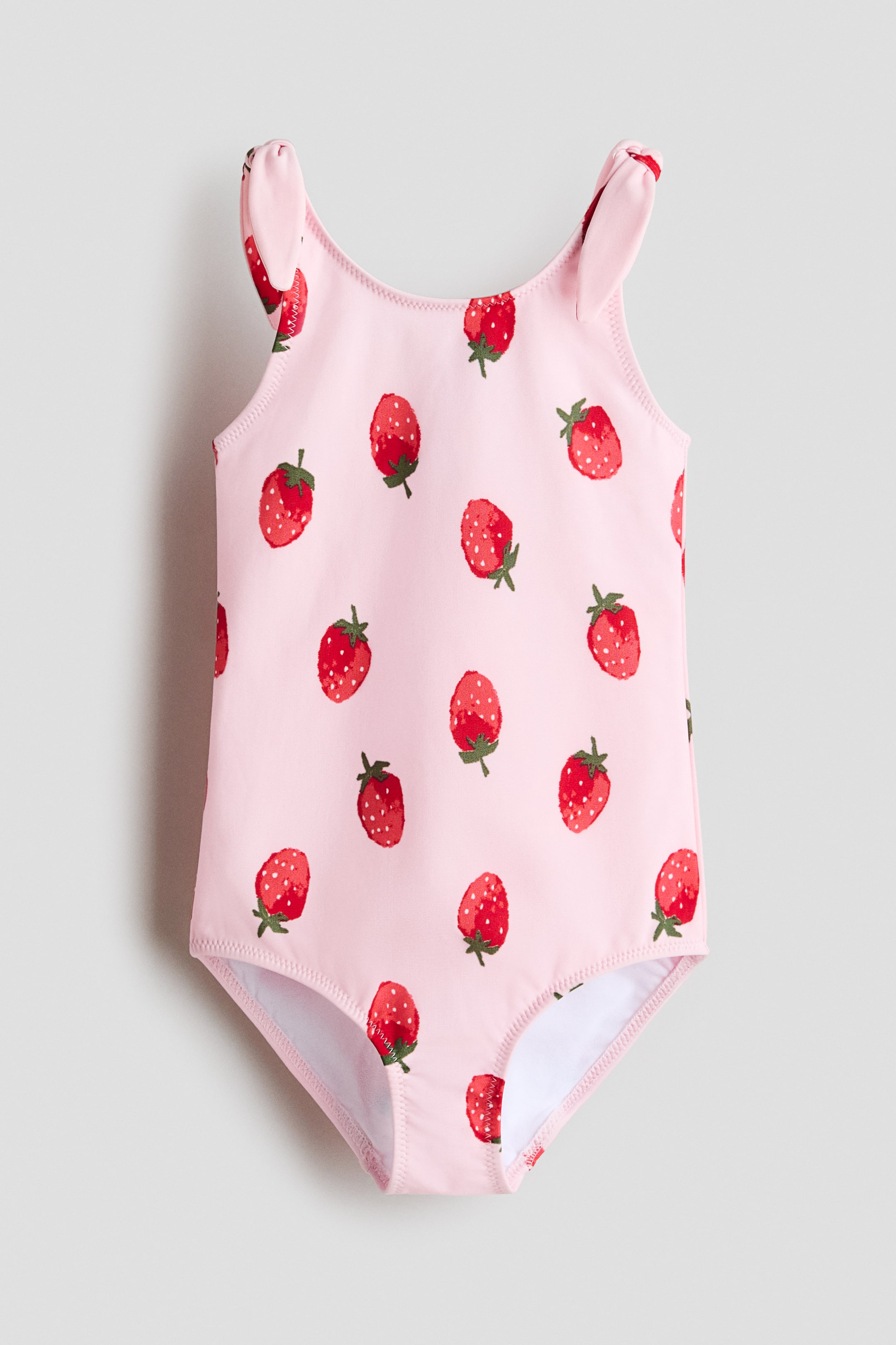 Prikaži veću sliku: Kupaći kostim je prikazan sprijeda, sa ružičastom osnovom i printom jagoda po celoj površini, a naramenice formiraju dekorativne čvorove. Takođe ima postavu.